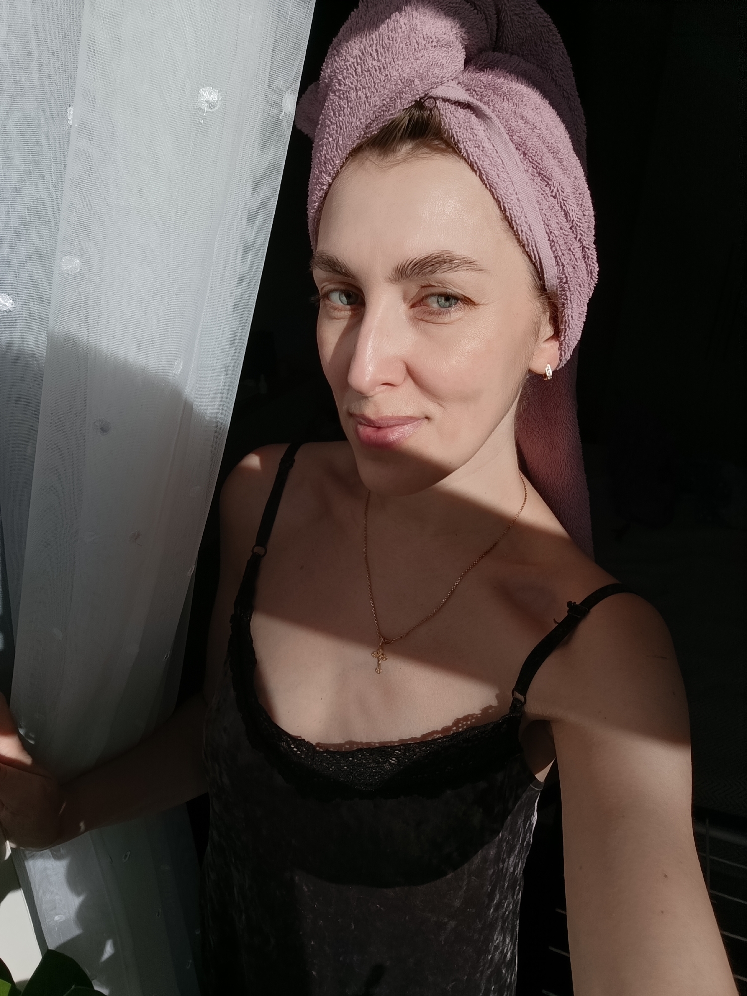A fair-skinned young woman poses indoors, likely in Chaikovsky, Russia given the request, with a lavender towel wrapped around her head and a gold cross necklace. She is wearing a dark-colored satin tank top with black lace trim. She stands next to a white curtain, her left hand holding the edge. Strong sunlight illuminates parts of her face, hair towel, and right arm, creating deep shadows on the right side of the frame and behind her. The background is dark and indistinct.