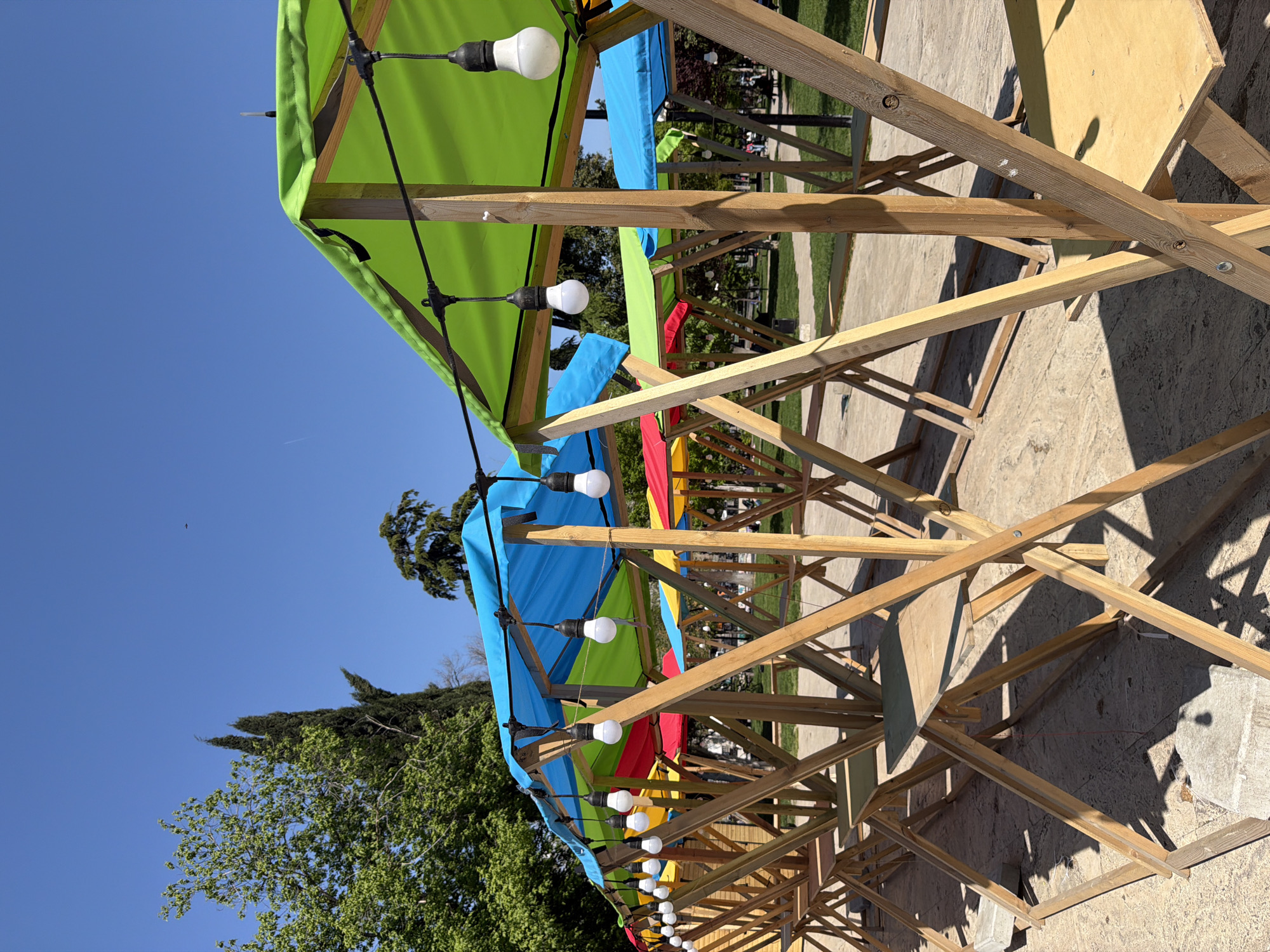 This media file depicts an outdoor scene featuring multiple wooden structures, likely market stalls or an outdoor dining area, under a clear blue sky. Each structure is covered with a vibrant, colorful awning, alternating between green, blue, red, and yellow fabrics.
Strings of white light bulbs are suspended beneath the awnings, suggesting the area is designed for both daytime and evening use, creating a festive ambiance. The wooden frames are visibly constructed with beams and supports, casting sharp, distinct shadows on the light-colored, paved ground, indicating bright sunshine.
In the background, lush green trees and foliage are prominent, adding to the natural outdoor setting. The sky is a deep, unblemished blue, with hints of a distant airplane or bird visible. No specific activity involving people is taking place, giving the scene a quiet, possibly pre-event or post-event atmosphere.
The time of day appears to be mid-morning or afternoon due to the strong shadows and clear sky. The weather is sunny and cloudless. The overall impression is one of a cheerful and inviting outdoor space, potentially a market or a cafe area in a park or public square in Tbilisi, Georgia. No visible text is present in the image.