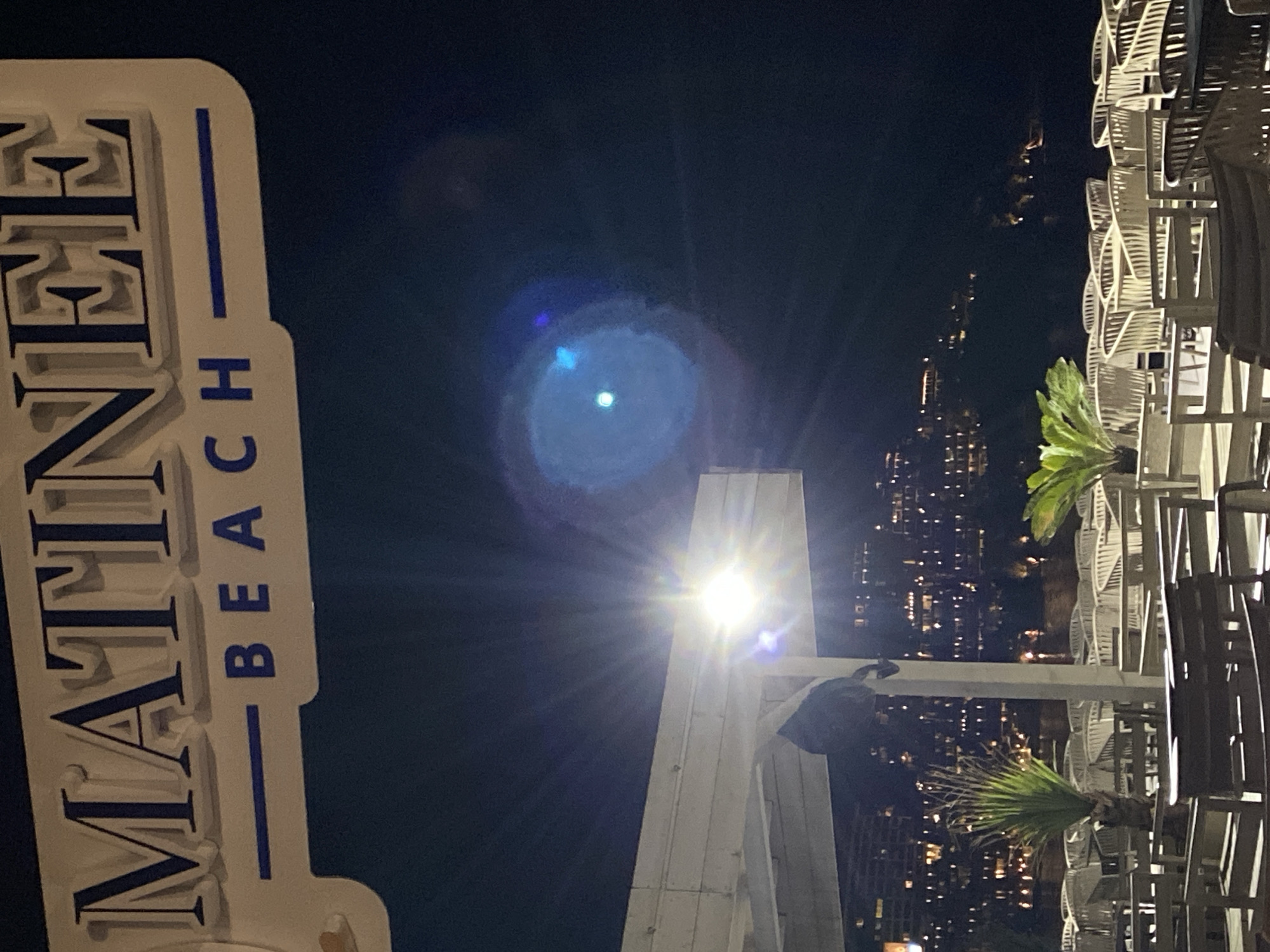The image depicts an outdoor establishment identified by a prominent overhead sign reading "MATINEE BEACH" in large, raised blue lettering on a white background. The scene is illuminated by artificial light at night. A bright light fixture is mounted on a white, horizontally planked overhead structure, generating significant lens flare. Below this structure, numerous light-colored, possibly woven, chairs are arranged around dark-colored tables, set on a wooden deck or platform. The establishment appears unoccupied by patrons. Palm-like plants are strategically placed among the tables. In the background, beyond the seating area, a dark body of water is visible, with the illuminated city lights of buildings on distant hills or across a bay reflecting on its surface. This setting is consistent with a beach club or restaurant located along the coast in Budva, Montenegro.