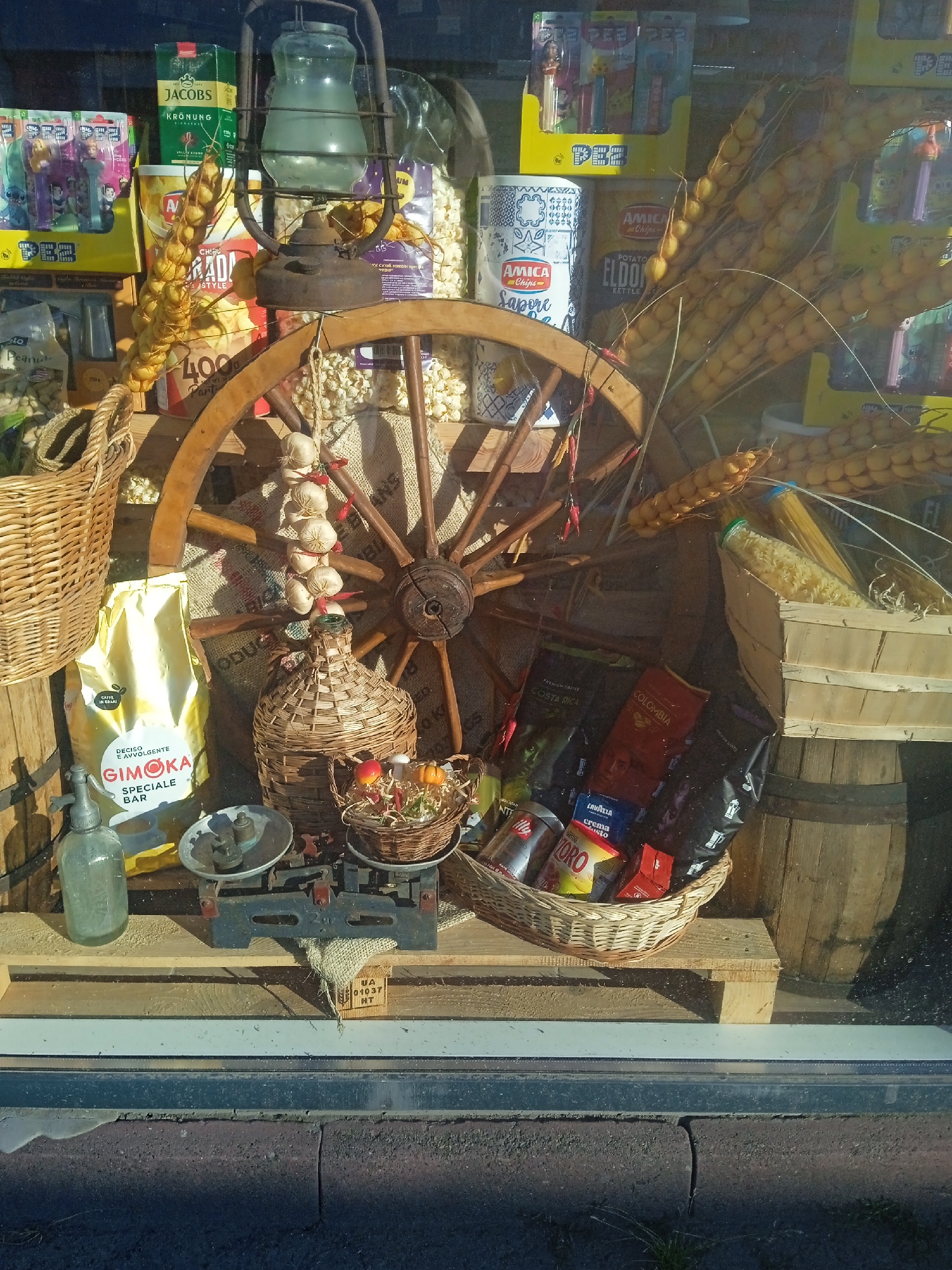The image displays a rustic window display, likely from a shop in Chernivtsi, Ukraine. The scene is rich with a variety of goods, suggesting a specialty food store or market stall. Dominating the center is a large wooden wagon wheel, its spokes weathered and aged.
Hanging from the wheel and surrounding structures are strings of dried garlic bulbs, their pale skins giving a sense of harvest abundance. Adjacent to the wheel, a large wicker basket holds a collection of coffee bags, prominently featuring brands like Lavazza, Costa Rica, and Colombia, indicating a focus on gourmet coffee.
Other items include a vintage-looking oil lantern with a glass reservoir, a woven bottle encased in wicker, and a small, rustic basket filled with decorative autumn produce like miniature pumpkins and apples. A classic mechanical scale, with weights placed on one of its pans, sits on a wooden surface. To the left, a yellow bag with Italian text, possibly flour or another baking ingredient, stands next to a glass seltzer bottle with a metal pump.
In the background, shelves are stocked with various packaged goods, including popcorn, potato chips, and colorful boxes of PEZ candy dispensers, adding a touch of consumer product variety to the overall rustic aesthetic. The display is set against a backdrop of burlap sacks, reinforcing the theme of natural and traditional products. The lighting suggests an outdoor setting, with sunlight casting shadows and highlighting the textures of the various objects. The overall impression is one of an artisanal and perhaps nostalgic presentation of food and drink items.