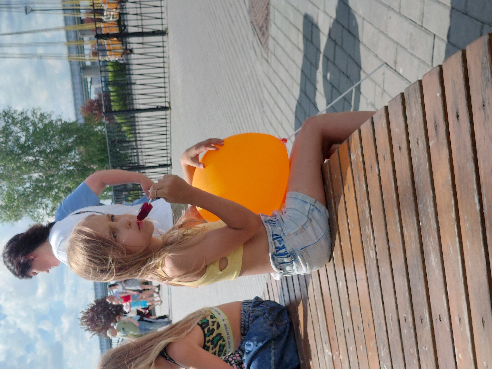The image captures a sunny outdoor scene, likely in Novosibirsk, Russia, judging by the context. In the foreground, a young girl with blonde hair is seated on a wooden bench, holding an orange balloon and a popsicle. She wears a yellow crop top and denim shorts. Her gaze is directed slightly away from the camera, with a thoughtful expression. Behind her, another person is partially visible, wearing a blue t-shirt and a white cap.
In the background, a children's amusement ride, possibly a swing ride, is visible with its chairs suspended in the air. The ground is paved with light-colored bricks, and shadows suggest it is daytime. Trees and greenery are present, indicating a park or recreational area. Further in the distance, other people can be seen, and there's a glimpse of a structure that might be a bridge or overpass. The sky is partly cloudy. The overall mood seems relaxed and summery. No visible text is present in the image.