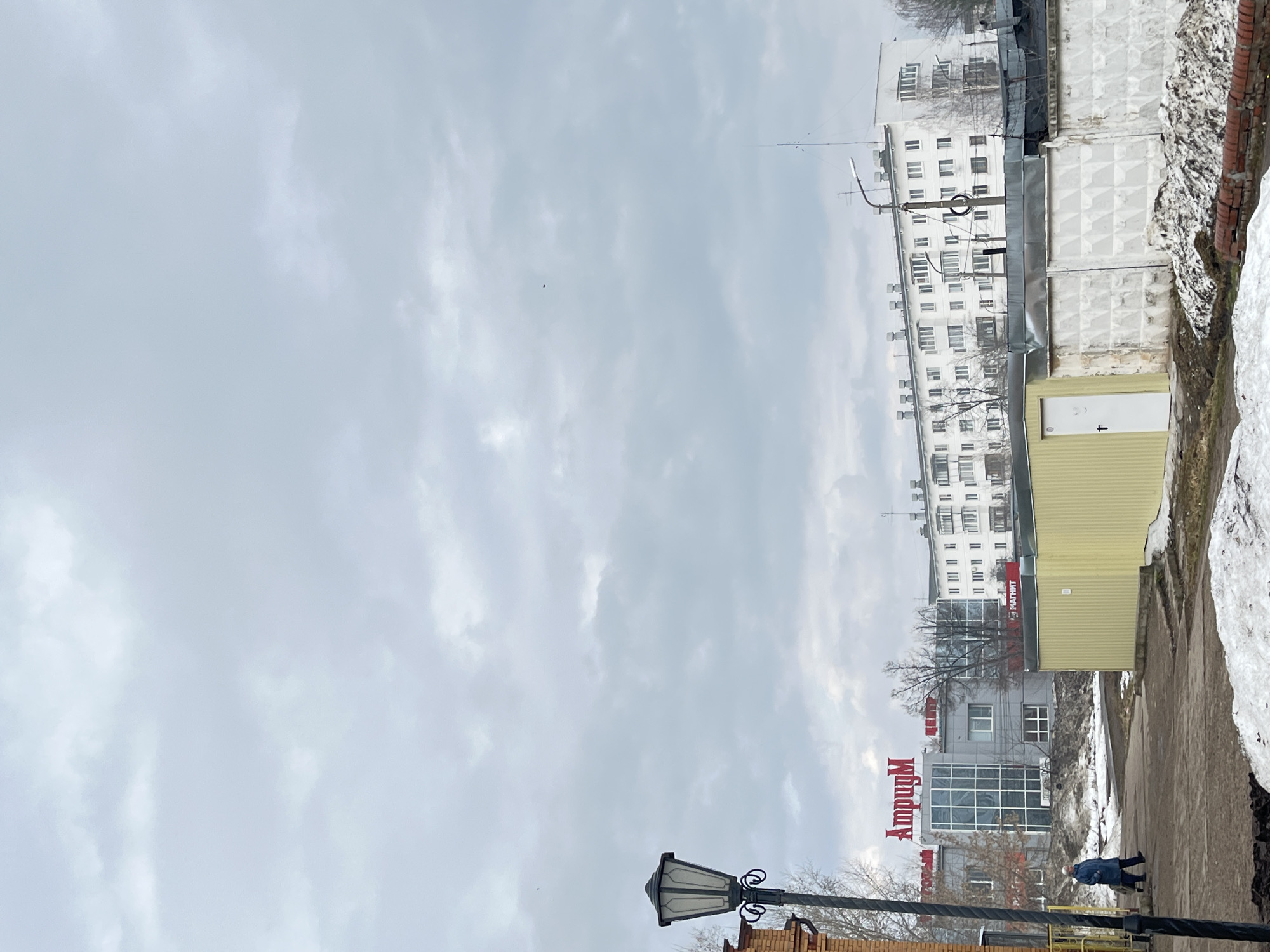 The image depicts a street scene in what appears to be a residential area of Chaikovsky, Russia, judging by the Cyrillic signage visible. The sky is overcast with a general gray tone. The focal point on the right is a multi-story building, possibly an apartment complex, painted in white with multiple windows. Adjacent to it is a rectangular structure, possibly a shop or storage unit, painted in a dull yellow color. To the left of this is a modern building with glass walls and a red sign reading "Ампудплх". A pedestrian is walking on the sidewalk in front of these buildings, facing away from the camera. The ground is a mix of concrete sidewalk and patches of snow, suggesting it is likely late winter or early spring. A streetlight is positioned at the bottom, left of the frame, suggesting it's part of the street infrastructure.