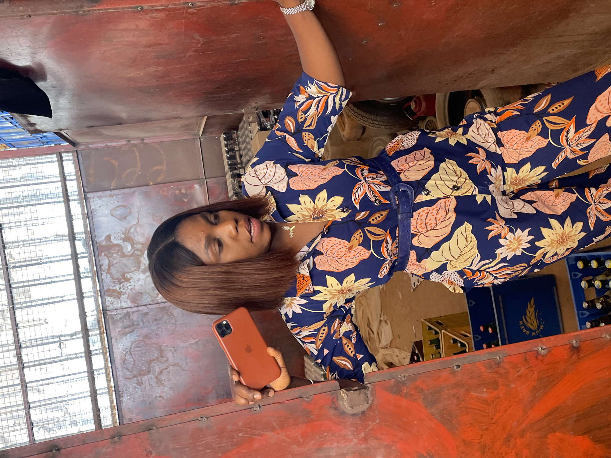 A young woman with brown hair stands inside a vehicle in Gura Top, Nigeria. She is holding an orange iPhone in her left hand and wearing a floral-print jumpsuit with a matching belt, and a silver watch on her raised right wrist. Her posture suggests she may be taking a selfie. The vehicle interior appears to be metal, with red paint on the visible surfaces. Crates of bottled beverages can be seen to her right, suggesting possible transport of goods. Behind her are glimpses of tires and various other items, indicating a somewhat cluttered interior.