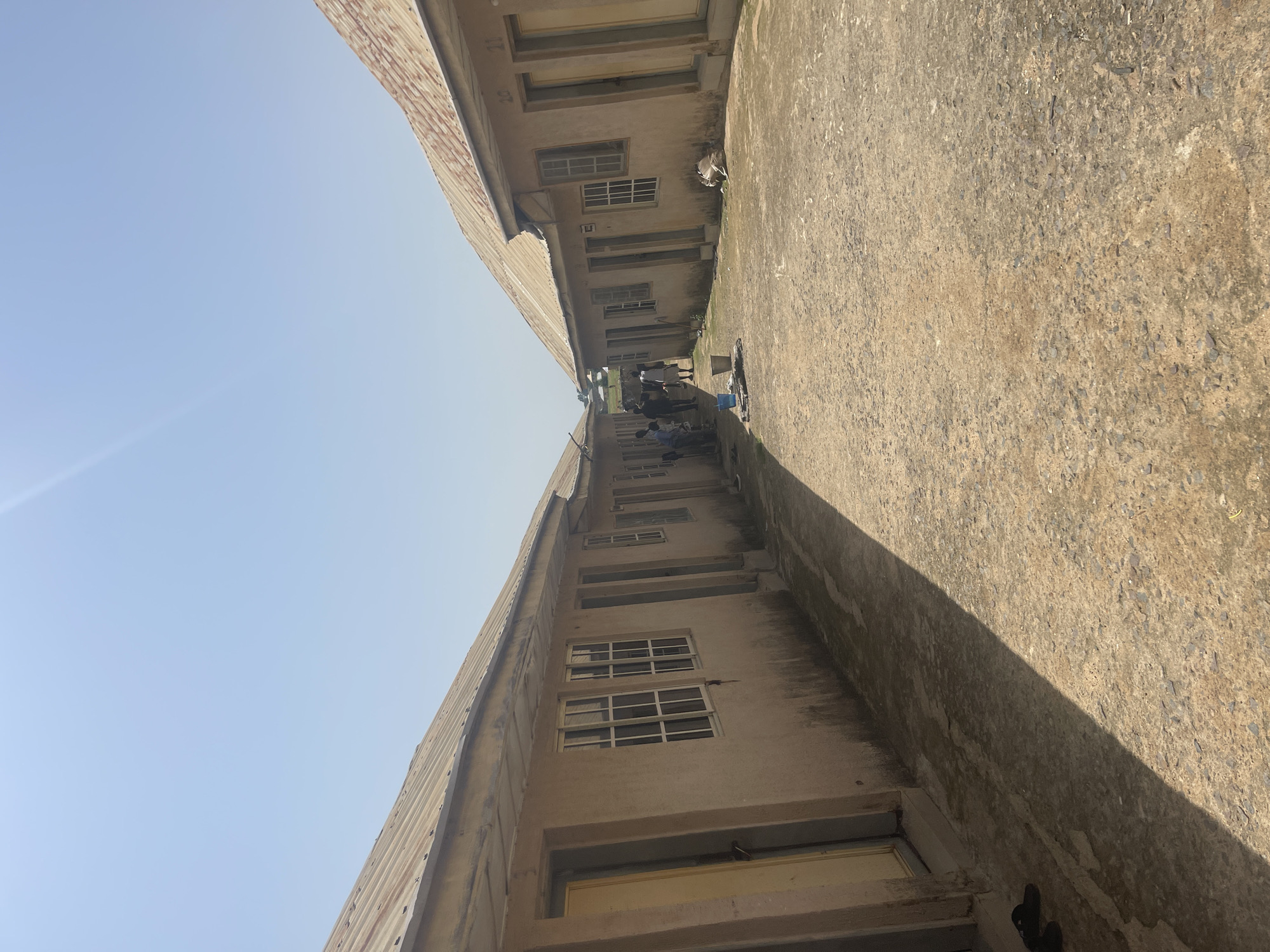 The image shows a view of an outdoor hallway between two buildings. The buildings appear to be made of concrete with corrugated metal roofs. There are several windows visible on the walls of the buildings. On the ground, there is a concrete walkway or courtyard.
Several people are visible in the hallway, appearing to be walking or standing around. One person is seen with a blue bucket. The scene is outdoors, with a clear blue sky visible above the buildings. The sun is casting a prominent shadow across the concrete surface, suggesting it is daytime, likely mid-morning or afternoon. The overall impression is a functional, perhaps institutional, setting.
There are no clear indications of specific emotions or a particular event taking place, beyond people being present in the hallway. The location is identified as Rafin Par, Nigeria. No visible text is discernable in the image.