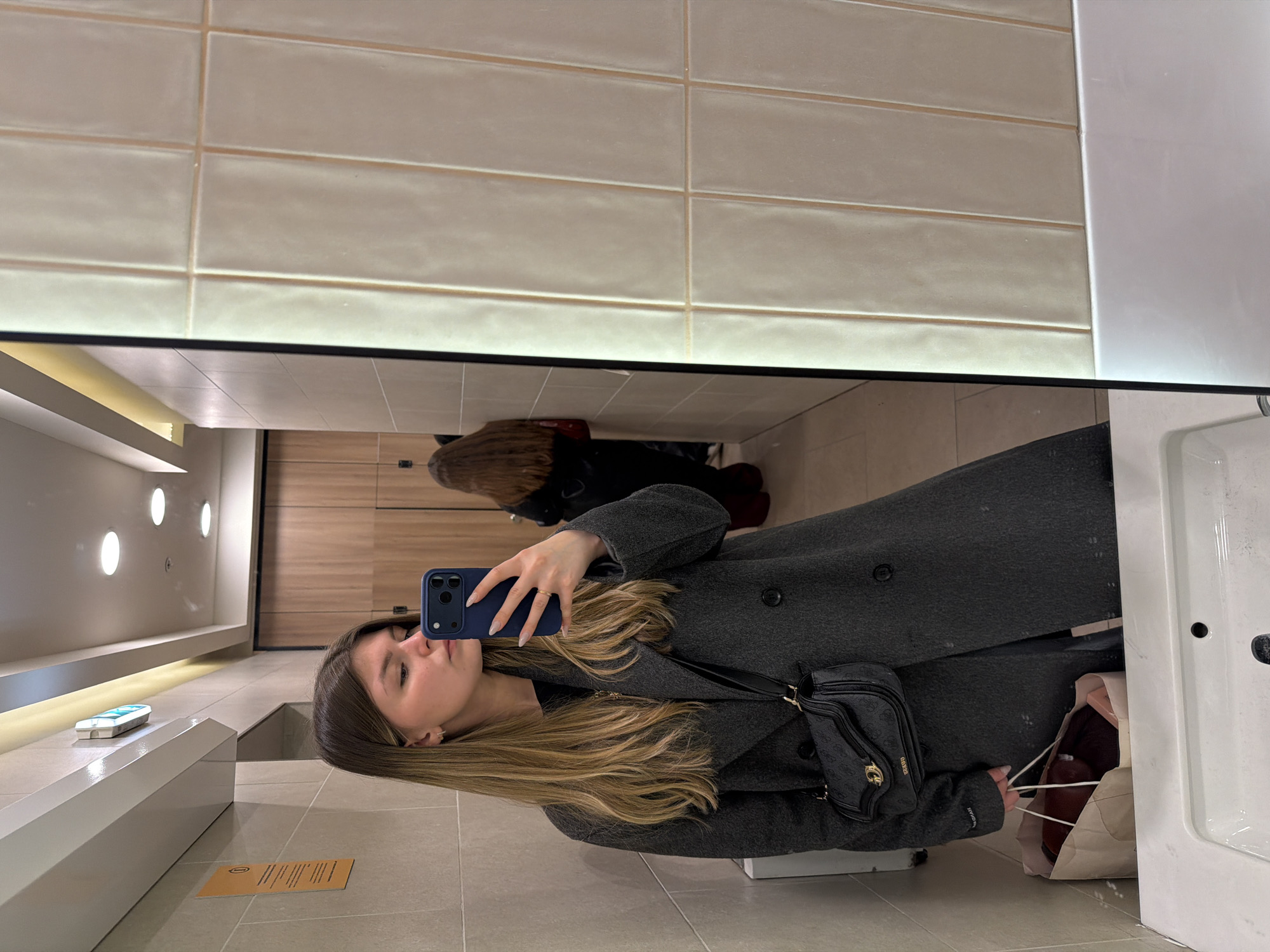 A young woman is taking a selfie in a restroom. The restroom has light beige brick-like wall tiles, a white counter with a white sink, and a long shelf with recessed lighting above. Other people are reflected in the mirror. The woman is wearing a long gray coat and a black purse. She is holding a blue smartphone in her hand. There is a shopping bag next to her. The location is potentially in the city of Saint Petersburg, Russia, but this cannot be definitively confirmed from the image.