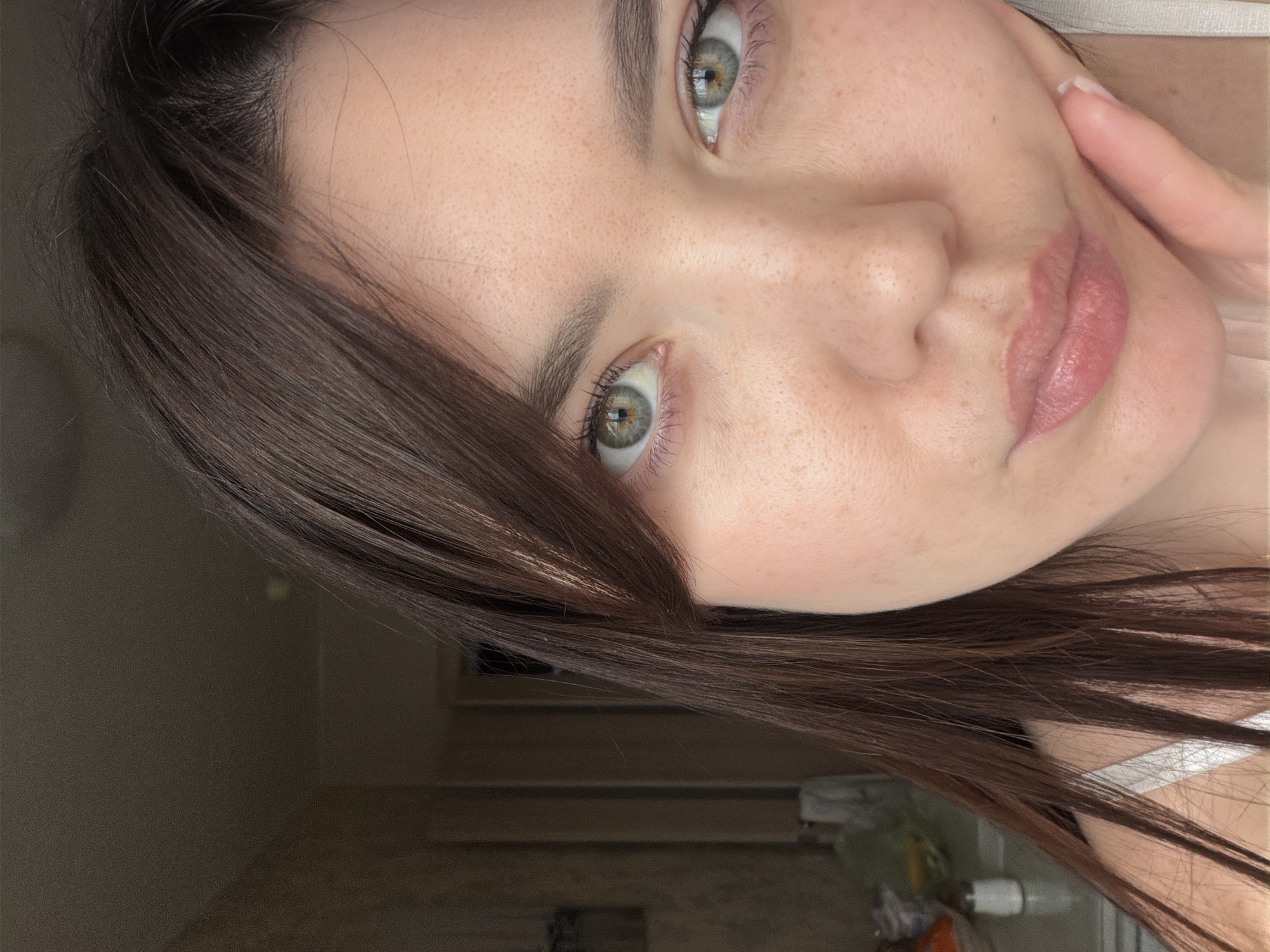 A close-up shot of a young woman with light skin and freckles. Her hair is dark brown and partially obscures her face. She has light green eyes. One hand is visible, touching her face. The background is blurred, but suggests an indoor setting with hints of a door and some objects in the lower right corner. No specific location within Chaikovsky, Russia can be determined from the image.