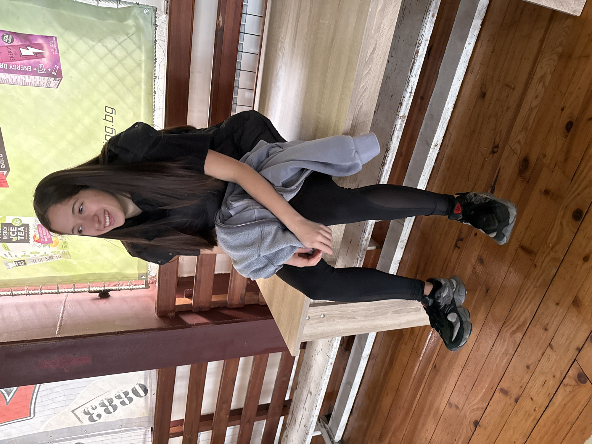 A young female, appearing to be in her late teens or early twenties, is seated indoors on a light-colored wooden bench. She has long, straight dark brown hair and a fair complexion, smiling and looking towards the left of the frame. She is dressed in a black short-sleeved top, a black sleeveless vest or jacket, black leggings with sheer panels on the sides, and black and gray athletic shoes. A light gray zippered hooded sweatshirt is held in her lap.
The environment features a polished light brown wooden floor. Behind the bench, there are dark brown wooden slats forming part of a wall or another seating structure. A dark reddish-brown vertical beam is visible on the left. Two large banners are affixed to the wall in the background. The right banner is light green and advertises "INSTICK ICE TEA" and an "ENERGY DRINK" with "ACAI flavour," displaying a partial URL ending in ".bg." The left banner is partially visible, featuring a red design and the numerical sequence "08830." A white radiator is discernible behind the wooden slats on the right side of the background. The presence of a ".bg" internet domain on the advertising banner indicates the location is in Bulgaria, specifically inferred to be within the city of Sofia. The setting appears to be a casual public indoor space, possibly a sports facility or youth center.