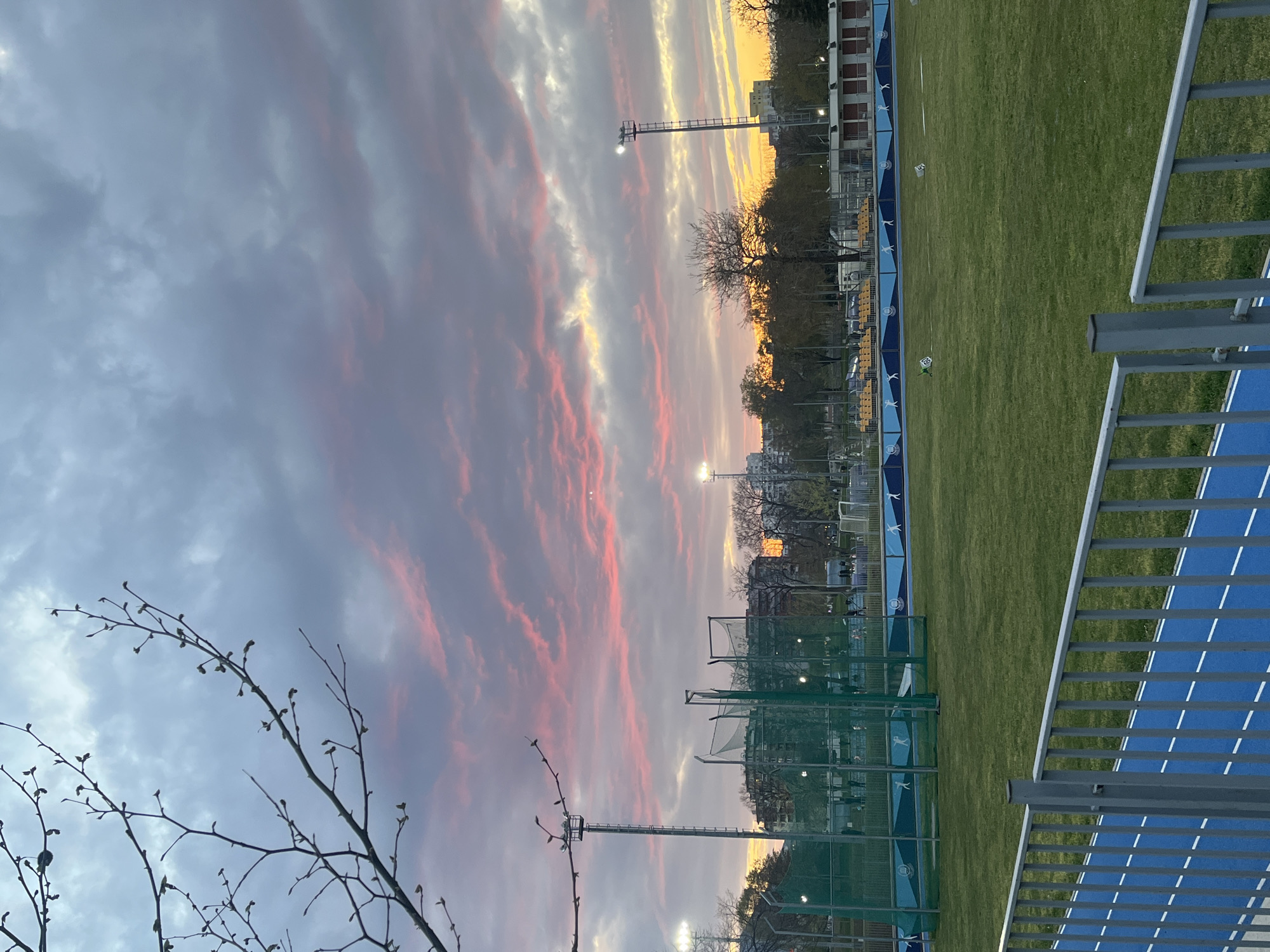 The image, captured in Sofia, Bulgaria, presents an outdoor sports complex under a dramatic, colorful sky, viewed in a rotated, vertical orientation. The top-left and central-left portions of the frame are dominated by an expansive sky, filled with dynamic cloud formations streaked with vibrant pink, orange, and yellow, mixed with softer grey tones, strongly suggesting either sunrise or sunset.
A vertical line roughly in the center divides the sky from the lower ground elements and forms the distant horizon. Along this horizon stand numerous trees, many of which are bare-branched with visible early buds, indicating late winter or early spring. Behind the trees, distant urban buildings are faintly visible. Several tall floodlight poles with their lights illuminated punctuate this middle ground.
The right side and bottom-right of the image showcase elements of the sports facility. A large expanse of bright green grass, possibly a football pitch or athletic field, is prominent. Adjacent to the grass, a bright blue track or pathway with white dashed lines runs parallel. A sturdy grey metal railing occupies the immediate foreground on the right, suggesting the image was taken from an elevated position beside the track. Further into the complex, green netting, additional fences, and yellow benches or bleachers are discernible. A small green marker cone with the number "19" printed on it rests on the grass. The blue boundary wall separating the grass from the track features repetitive white stylized figures, resembling athletes, and small, indistinct logos. The scene is devoid of people, conveying a tranquil moment at dusk or dawn.
