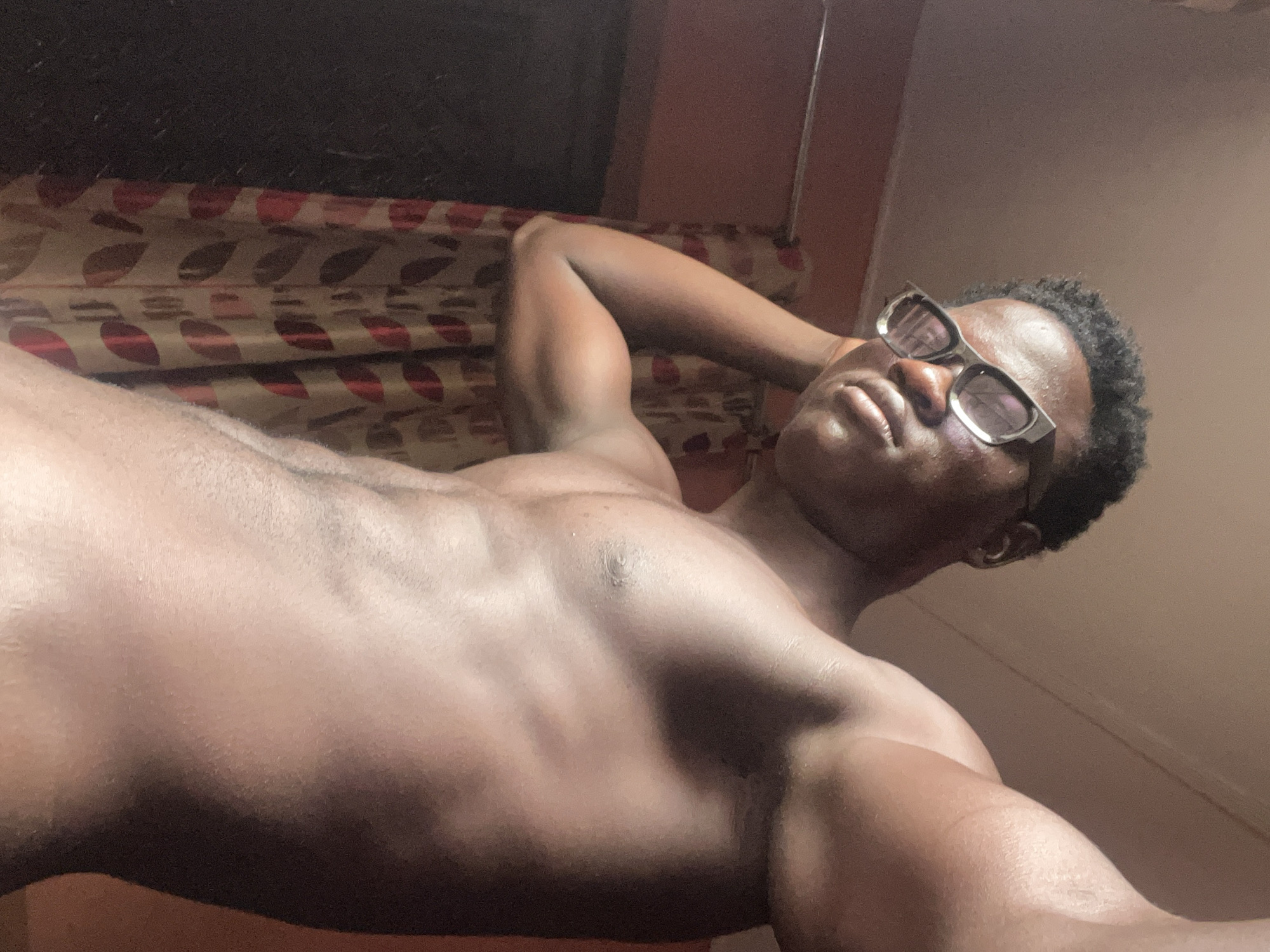 A shirtless, dark-skinned man with short, curly hair and wearing glasses is reclined on a bed. The man has his arms positioned behind his head. The background features a headboard with patterned upholstery and a plain wall. The image appears to be taken indoors. There is no evidence to suggest that this image was taken in Mosafejaw, Nigeria.