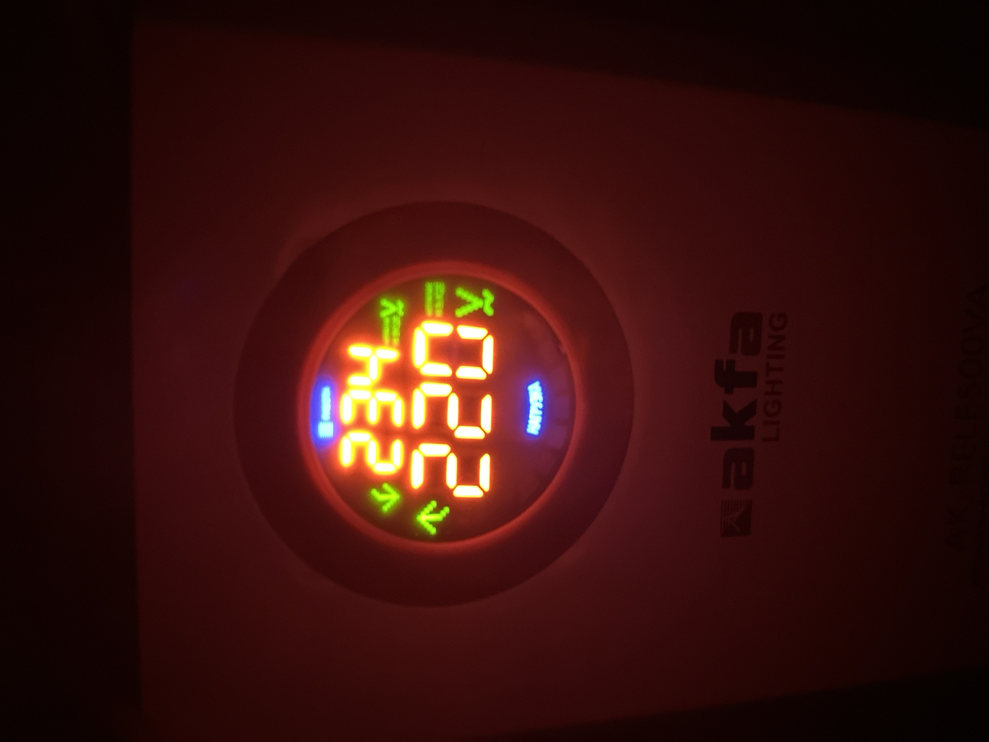 The image displays a close-up of a red electronic device, likely a voltage stabilizer or regulator, with a circular digital display. The display shows two sets of numbers in bright orange: "234" above and "220" below. To the left of these numbers are green upward and downward arrows, indicating potential adjustments or readings. Above the numbers, a blue label reads "НАГРУЗКА" (which means "LOAD" in Russian). Below the digital display, a logo with the text "akfa LIGHTING" is visible in a darker shade of red. The device appears to be indoors, given the lack of natural light and the artificial glow of the display. The background is dark and out of focus, suggesting the photo was taken in low light conditions. There are no people or specific city landmarks visible. The location of Fergana City, Uzbekistan is not depicted in the image.