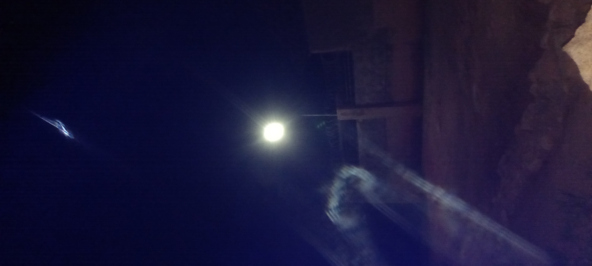 The image depicts a nighttime outdoor scene, likely in Kwang, Nigeria, illuminated by a single, bright street lamp. The sky is a deep, dark blue, with a faint, wispy streak of light visible in the upper portion. Below the sky, the street lamp is prominently positioned in the center, casting a strong, diffused glow.
In the middle ground, a low, dark wall with barbed wire along the top is visible. Adjacent to the wall, to the right of the street lamp, is a concrete pillar. To the left of the wall, in the foreground, there are blurry, streaking lights, possibly caused by motion blur or light reflection. The ground in the foreground appears to be rough and uneven, with some lighter patches that could indicate sand or loose stones. The overall atmosphere is dark and somewhat abstract due to the low light conditions and potential motion blur. There are no visible people or distinct actions taking place.