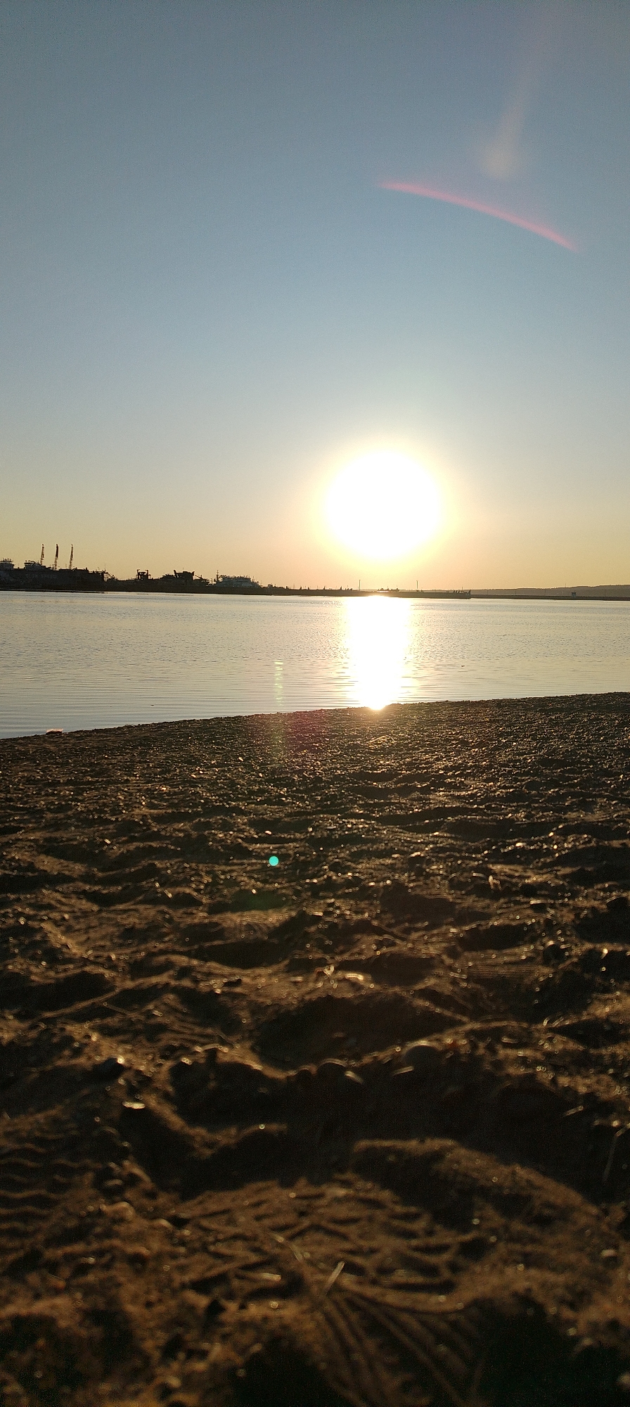 The image depicts a sunset over a body of water, likely a river or bay, from a low vantage point on a sandy or pebbly beach. The sun is a bright, circular orb positioned on the horizon, reflecting strongly on the water's surface. In the background, silhouetted structures or ships are visible along the shoreline. The sky is a gradient of light blue near the top to a warm, golden hue closer to the horizon. There is a slight lens flare visible in the upper part of the sky. The foreground shows the texture of the sand or pebbles on the beach. The city is unspecified and its location is unknown.