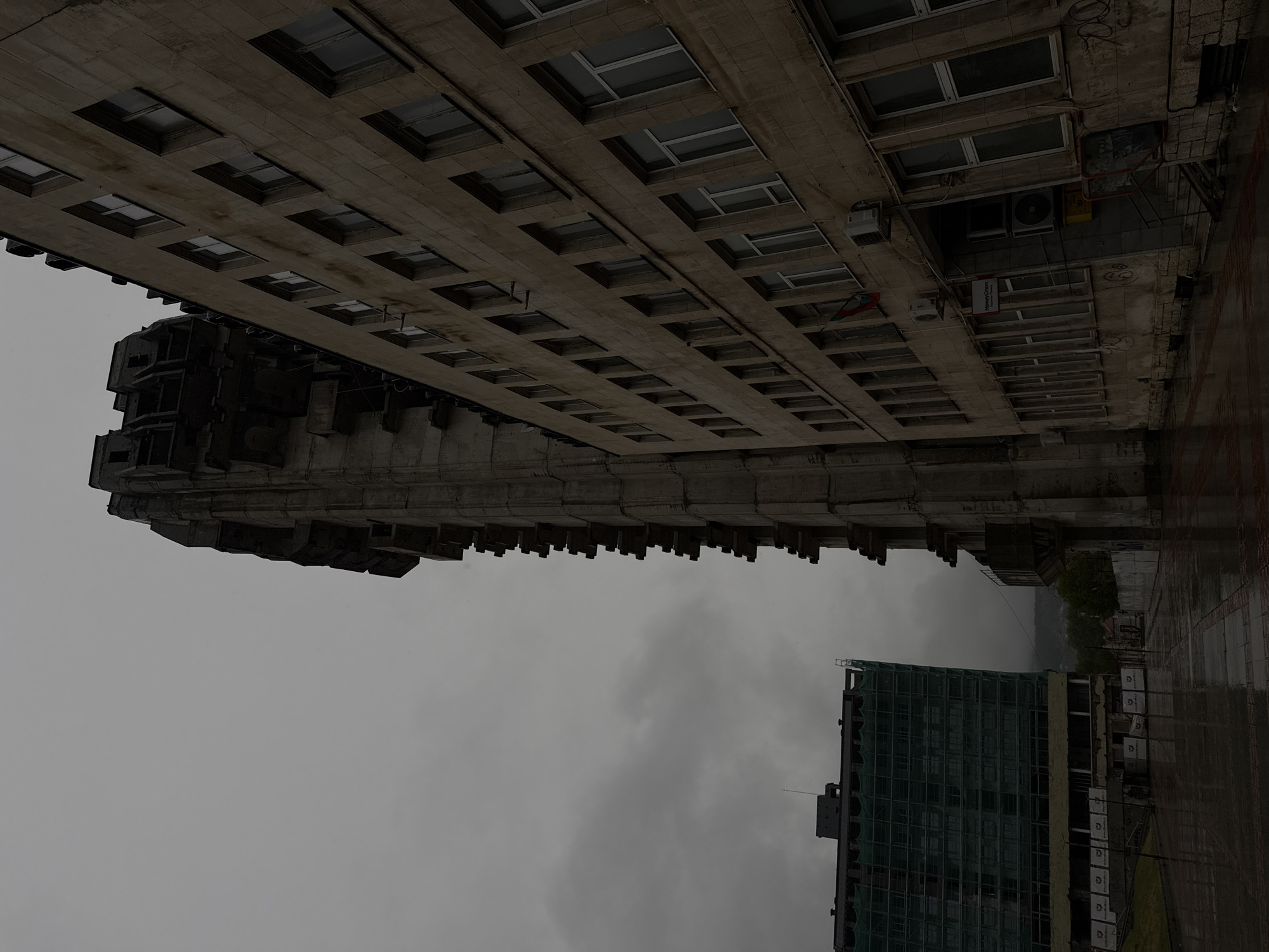 A vertical urban landscape photograph, taken on an overcast day, captures two distinct buildings in Shumen, Bulgaria. The image, rotated 90 degrees clockwise, depicts a scene looking upwards towards the sky.
On the left, a large, multi-story building dominates the frame. Its facade, constructed from concrete or stone, features numerous rows of rectangular windows. Towards the foreground of this building, a darker, imposing architectural section, possibly a tower or a unique structural element, is prominent. This section exhibits a series of angular, block-like protrusions, characteristic of Brutalist or monumental design. Visible on the building's facade are air conditioning units, various wires, and a red flag-like object. A rectangular sign with the text "MoneyGram" is discernible.
On the right side, further in the background, stands a modern building, partially obscured by green construction netting or scaffolding, indicating renovation or ongoing construction. White rectangular panels, some featuring a 'D' logo, are visible on its exterior.
The sky is uniformly grey and heavily overcast. The ground, visible in the lower right, appears wet and reflective, suggesting recent or ongoing rain. No people are visible, and the scene is static, portraying an atmospheric urban environment under grey skies.