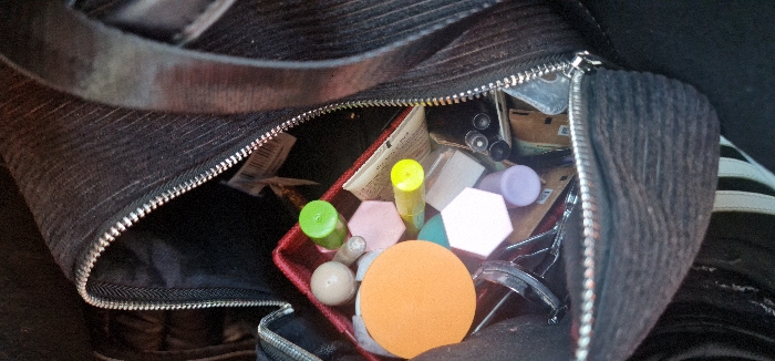 A close-up, top-down view depicts the partially unzipped interior of a dark, likely black, ribbed fabric bag, located in Cenon, France. The bag's zipper is silver-toned. Inside, a red rectangular container holds an assortment of personal items. These include an orange circular cosmetic sponge, two hexagonal sponges (one light pink, one white/light beige), and several cylindrical tubes with colored caps (one green, one yellow, one light purple). A light beige spherical object, a silver-toned eyelash curler, and multiple dark cylindrical objects resembling pens or brushes are also visible within the bag. Various crumpled papers or labels are interspersed among the items. The bag's interior lining is dark. A dark fabric strap is visible across the top, and a portion of a white-striped object is partially visible on the far right side of the bag's exterior. No individuals are present, and no actions are discernible.