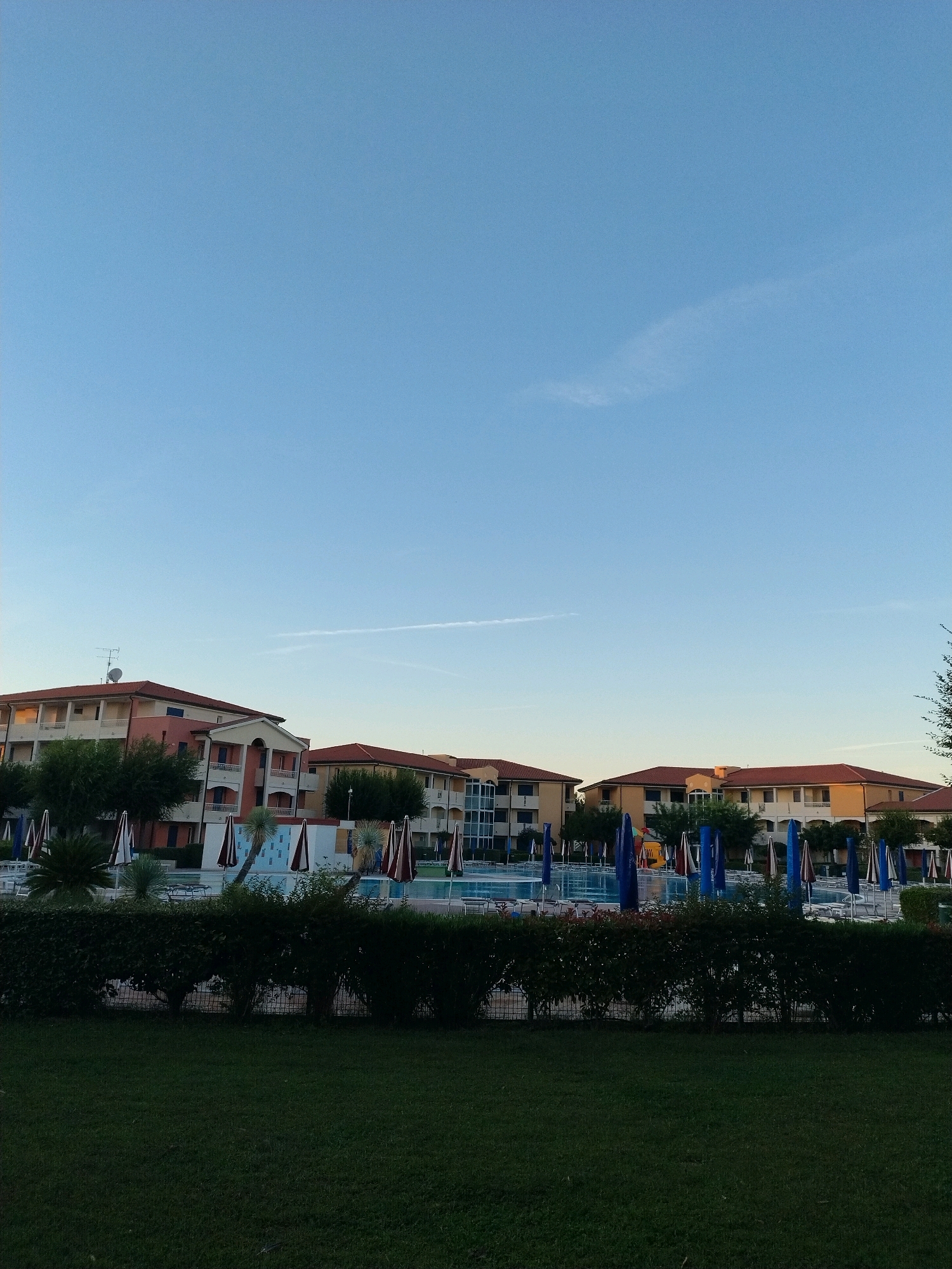 The image depicts a resort area in Caorle, Italy, likely during the early morning or late evening, given the soft light and clear sky. The scene is dominated by a large, blue swimming pool, partially visible in the middle ground, surrounded by lounge chairs and umbrellas. Several multi-story buildings with reddish-brown tiled roofs and cream-colored facades are situated behind the pool area, suggesting hotel or apartment accommodations.
A dense, dark green hedge runs across the foreground, separating a manicured lawn from the resort facilities. The lawn itself appears well-maintained and is a deep, rich green. The sky above is a clear, pale blue with a faint, wispy cloud or contrail visible in the distance. There are no people visible in the frame. The overall impression is one of a tranquil and well-kept vacation destination.
