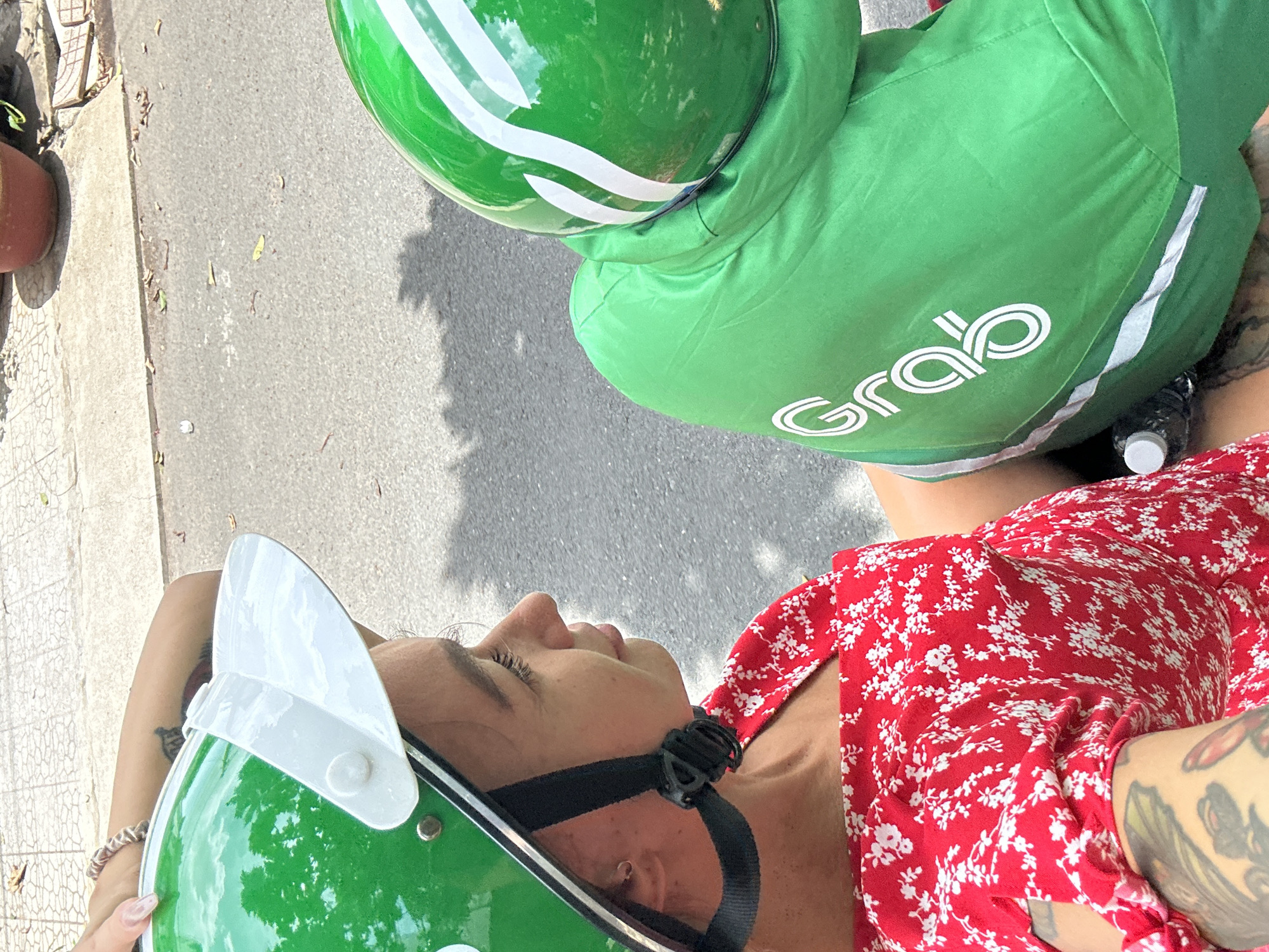 An overhead shot shows a woman in a red floral dress and a green helmet with a white visor, riding on a Grab motorcycle in Đồng Nai, Vietnam. The Grab driver, wearing a green helmet and green jacket with the Grab logo, is visible in the upper right of the frame. Pavement and a flower pot are visible to the left.