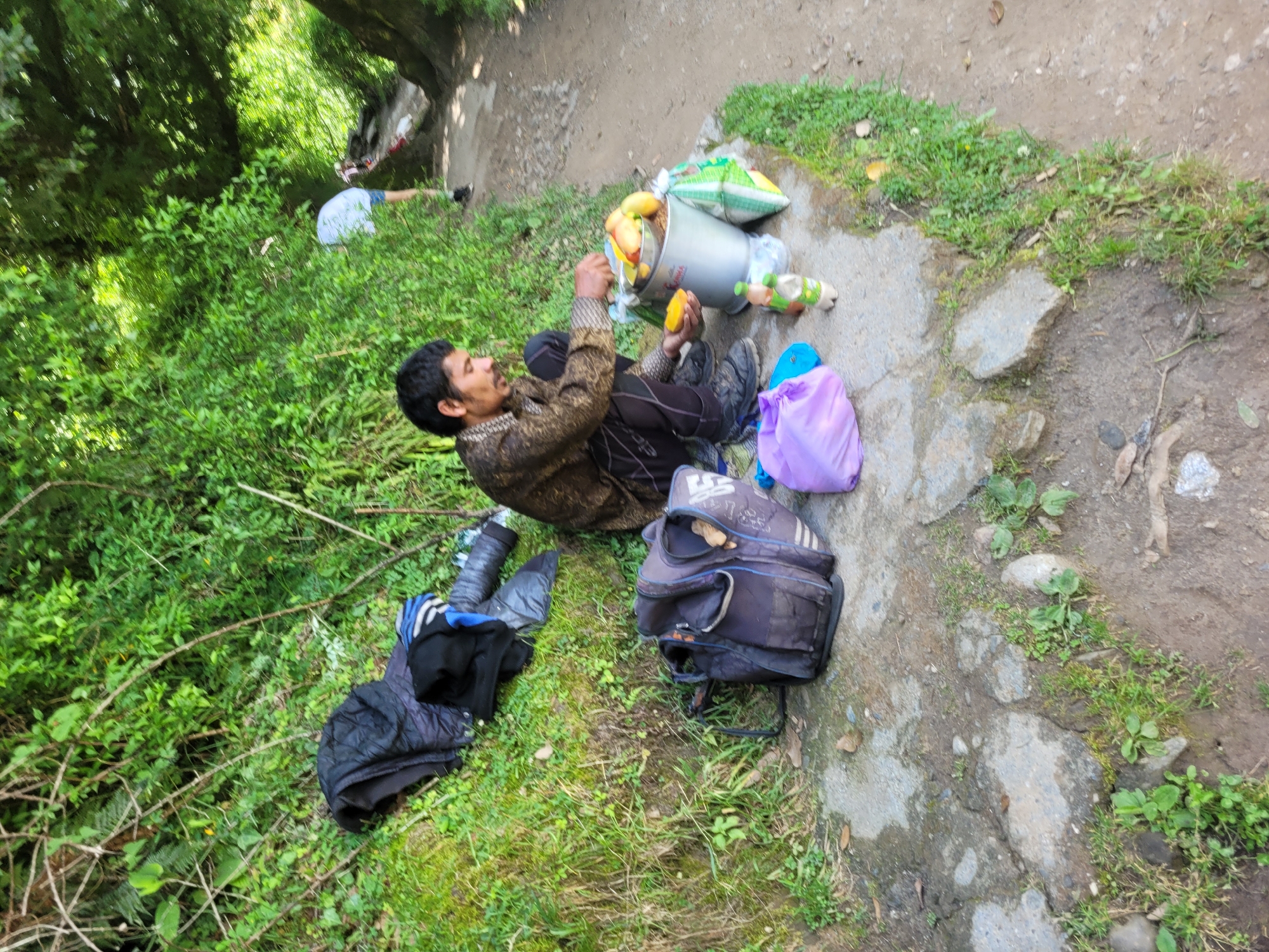 In Vashisht, India, a man is squatting on a stone path next to a grassy embankment. He is wearing a patterned brown shirt and dark pants. In front of him, on the path, is a metal bucket filled with what appears to be fruit, possibly mangoes. He is holding a piece of fruit in his right hand and a knife in his left, as if preparing to cut it. To his left, there are several bags, including a dark backpack and a bright purple bag. Behind him, a person wearing a light blue shirt and shorts is visible walking away on a dirt path. The environment is lush and green, with dense foliage surrounding the area.