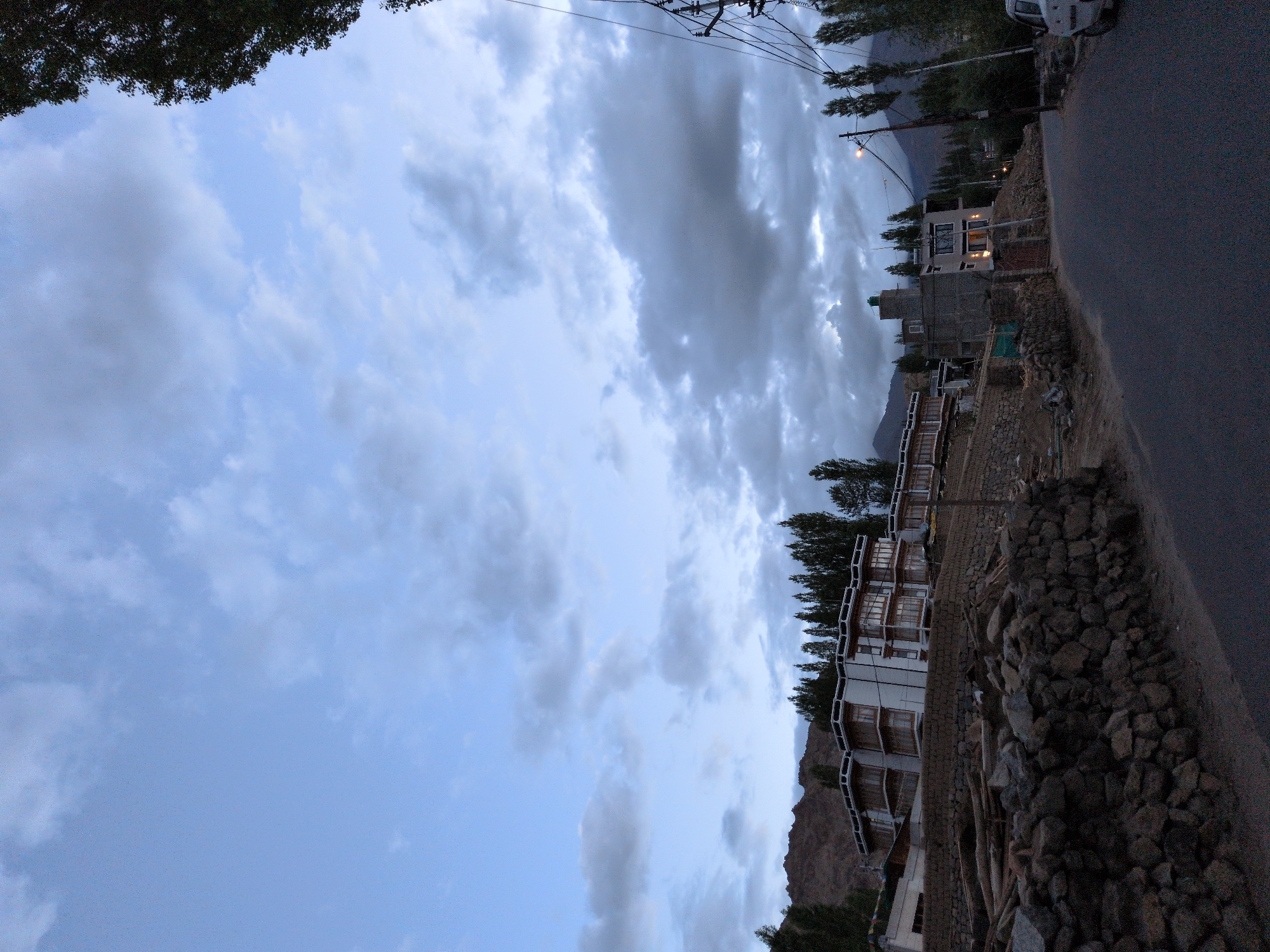 The image depicts a scene in the city of Leh, India, at dusk. A paved road curves through the foreground. On the left side of the road, a stone wall borders a terraced hillside. Above the wall, a large, multi-story building with a modern architectural style is visible, featuring white walls and numerous wooden-framed windows. Behind this building, a row of tall, slender trees stretches across the landscape, with darker, mountainous terrain in the background. On the right side of the road, a utility pole with wires stands tall, and further down, another building with illuminated windows is situated on a slope, with more trees and mountains beyond. A white car is parked on the right side of the road near the utility pole. The sky is filled with a mixture of light and dark clouds.