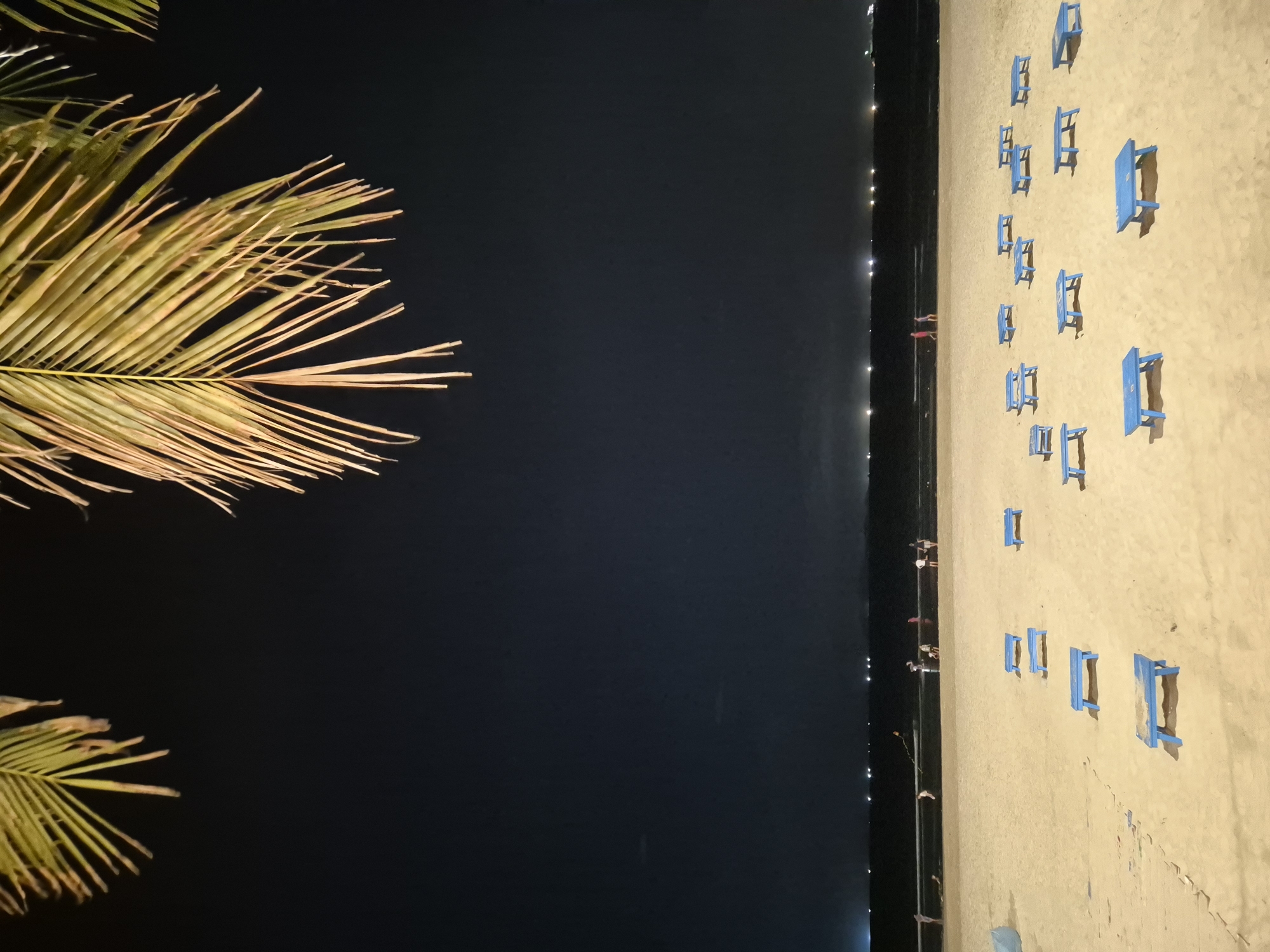 The image depicts a night scene on a sandy beach, likely in Đà Nẵng, Vietnam, with a cluster of blue picnic tables and benches scattered across the foreground. Palm fronds hang from the top of the frame, silhouetted against a dark night sky dotted with distant lights reflecting on the water. In the background, a calm sea meets the shore, and several people are visible near the waterline, some walking and others engaged in activities like flying a kite. The overall atmosphere is serene and tranquil, with the gentle glow of distant lights illuminating the beach and sea.