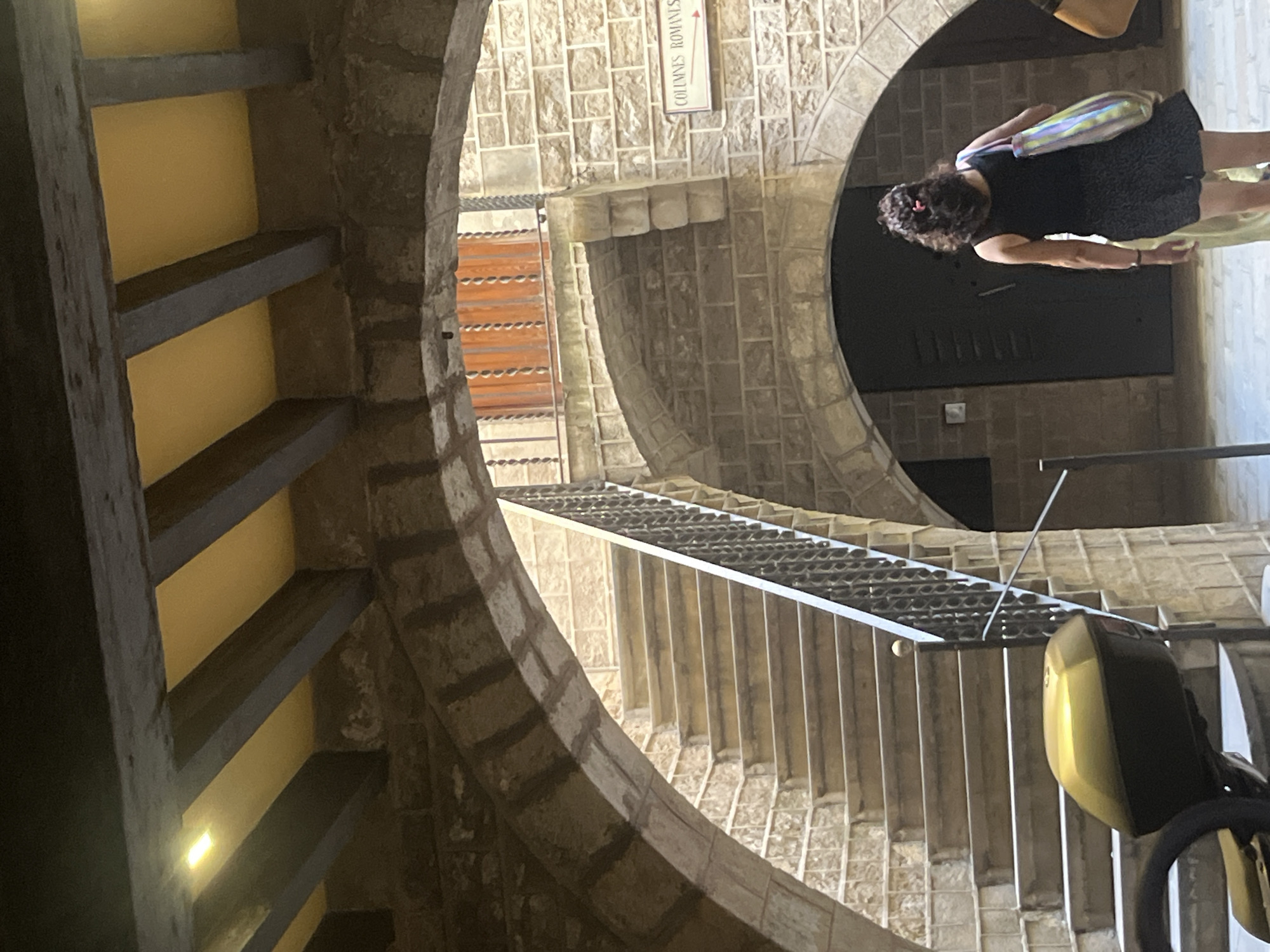 A low-angle shot shows the interior of a stone structure, possibly a tower or circular building, featuring spiral stairs. The structure appears old, with exposed stone and archways. A sign points to "COLUMNES ROMANES." A woman wearing a dark dress and carrying a colorful bag stands near one of the archways. The image captures the architectural details of the building and its internal layout, which has a medieval or historical feel. The shot is taken inside the Museu d'Història de Barcelona (MUHBA) located in Barcelona, Spain.