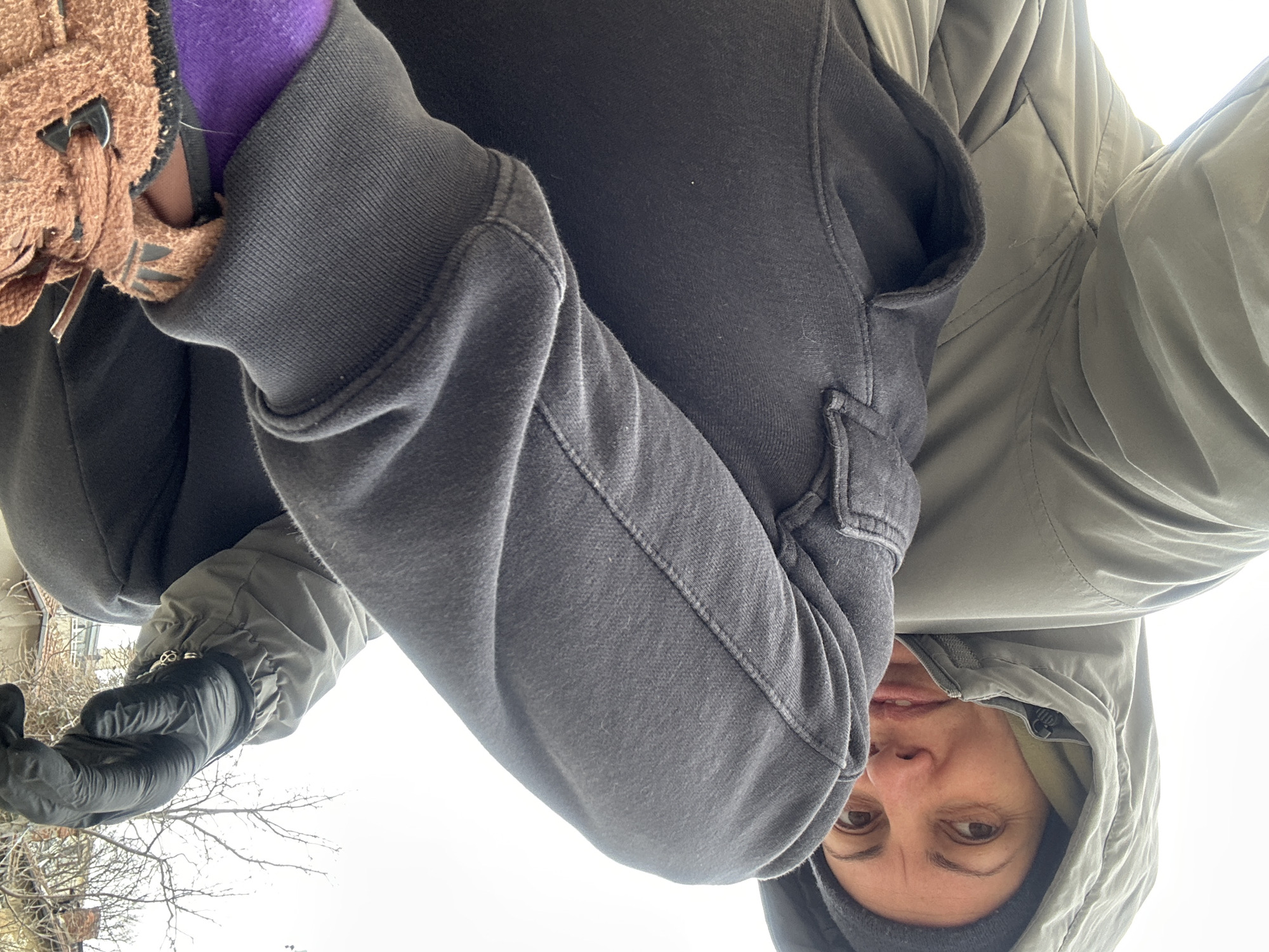 A person is holding a brown slipper, wearing black gloves, a purple sleeve, a grey sweatshirt, and a green hooded jacket. The person has brown eyes and is wearing a grey beanie. In the background, there are bare tree branches and a building. The scene is likely outside on a cloudy day. The location is Sofia, Bulgaria.