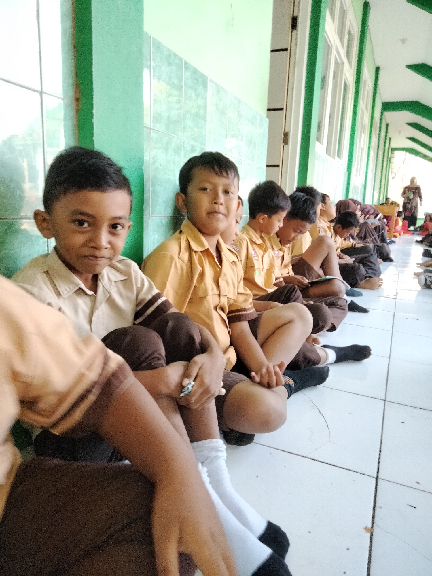 The image shows a group of elementary school-aged boys sitting on a tiled floor in a hallway. They are wearing light brown uniforms with dark brown trim and dark socks. One boy in the foreground is looking directly at the camera with his knees pulled up to his chest. Other boys are seen in the background, some looking at books or tablets. The hallway has light green walls and a white tiled floor. Sunlight streams in from the right, casting shadows. In the distance, a woman in a patterned dress and headscarf is visible near a desk. The scene appears to be at a school in Pucakwangi, Indonesia, likely during a lesson or break.