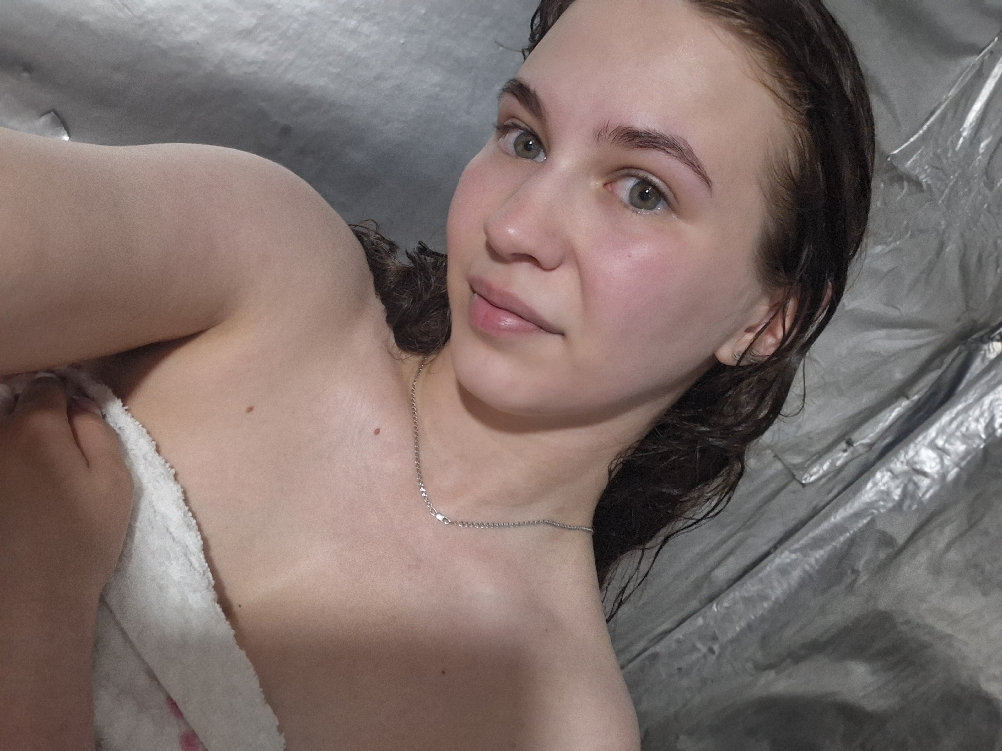 A young woman is pictured lying down on what appears to be a silver-colored tarp or sheet. She is partially covered by a white towel. She is looking directly at the camera with a neutral expression. She has fair skin, light brown hair that appears to be wet, and light-colored eyes. She is wearing a thin silver chain around her neck. The background is indistinct and appears to be the same silver-colored material as the tarp. The location Nizhnyaya Gar', Russia is not verifiable from the image.