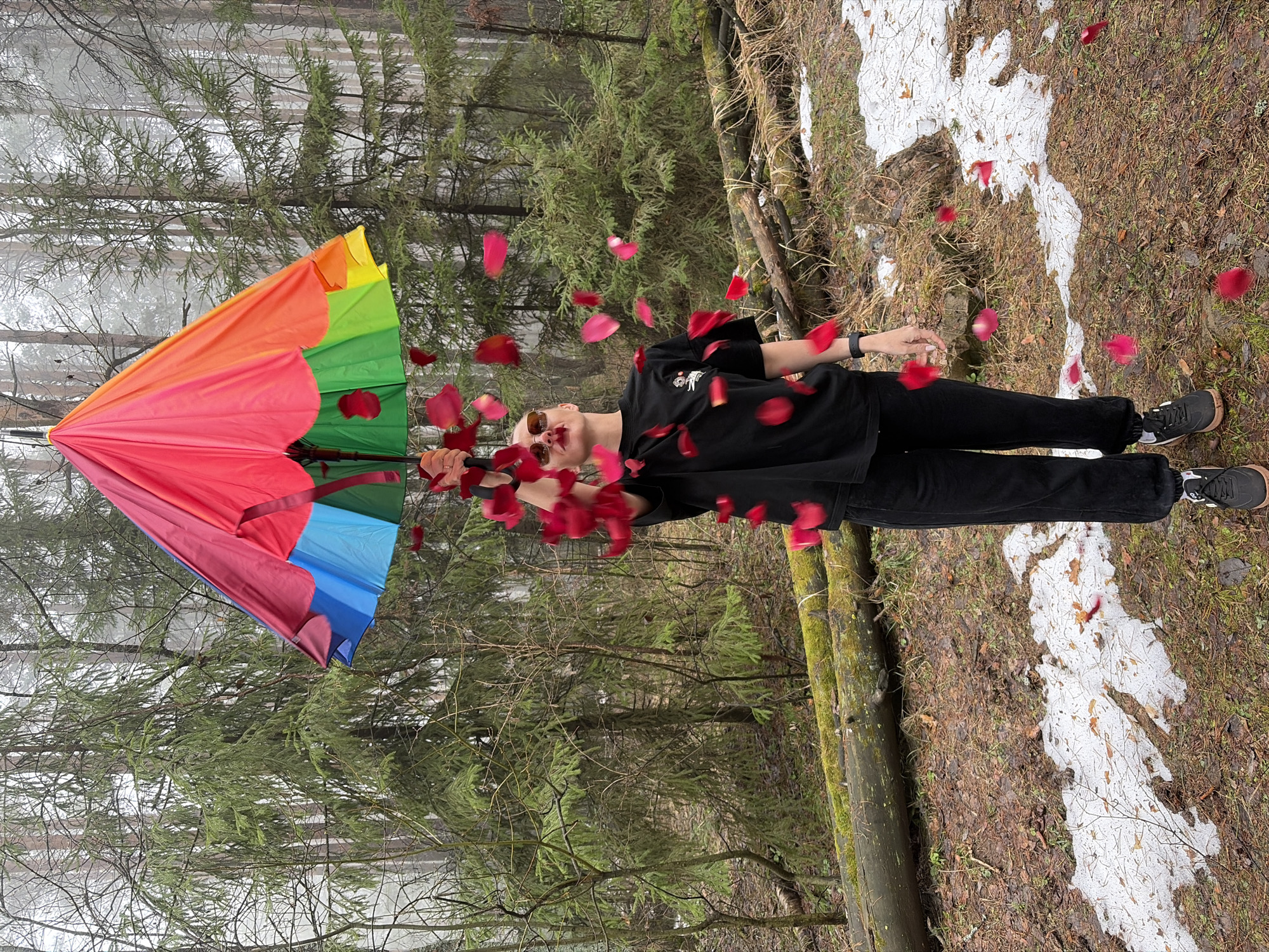 A person in black clothing and sunglasses is in a forest with an open, multi-colored umbrella. Red flower petals are scattered around the person. The ground is covered with brown leaves, green vegetation, and patches of snow. The location appears to be near Прикамский, Russia, as indicated by the landscape and vegetation. The person is positioned near a moss-covered log.