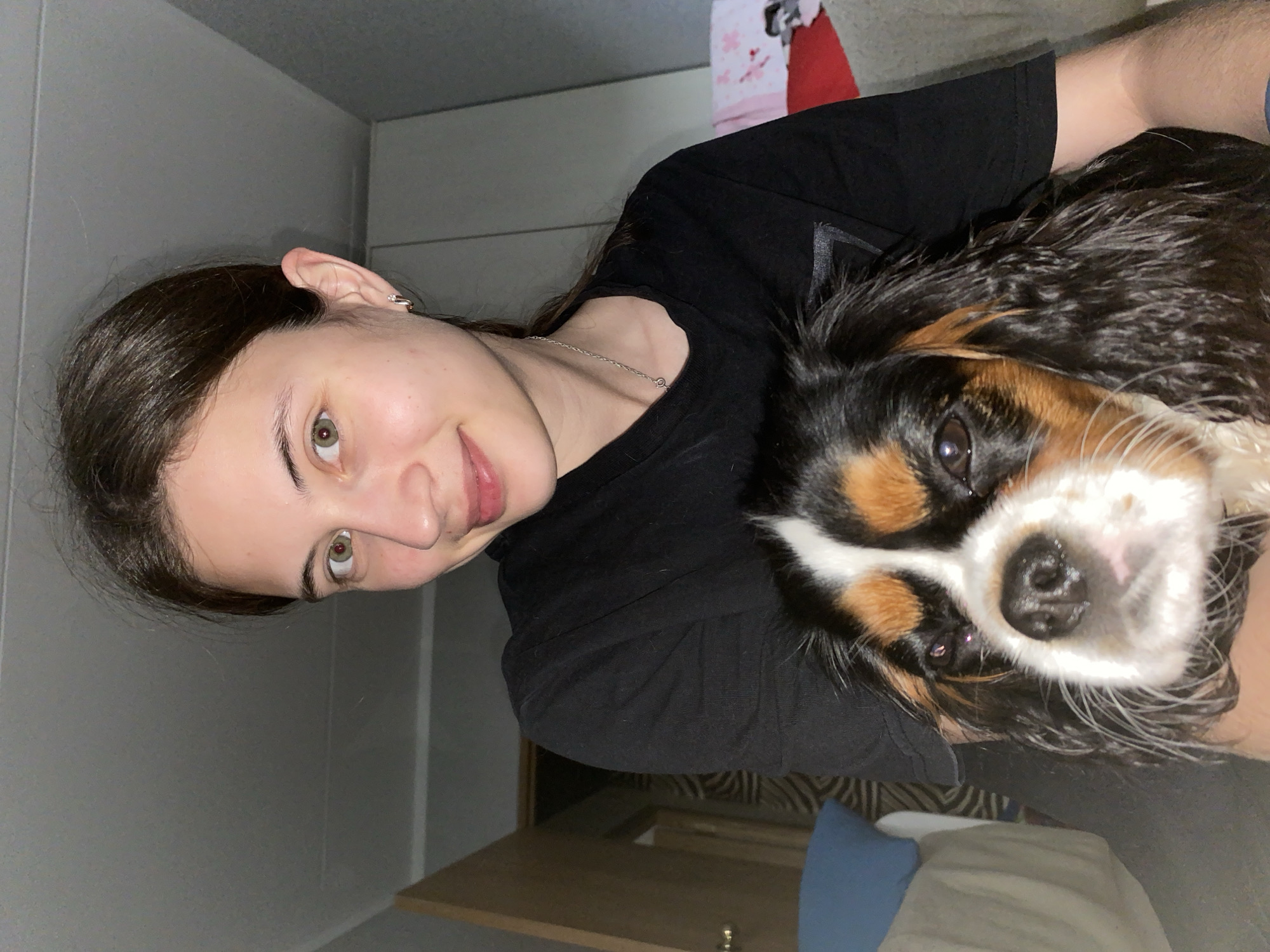 The image shows a young woman with dark hair and green eyes, wearing a black t-shirt. She is holding a dog, likely a Cavalier King Charles Spaniel, which is black, brown, and white. The dog's face is close to the camera, and its nose is prominent. The woman is smiling, and the dog appears to be looking at the camera. The background is dimly lit, suggesting an indoor setting, possibly a home. The overall mood is warm and affectionate, with the woman and her pet sharing a moment. The image seems to be taken in the evening or at night due to the lighting conditions. No text is visible in the image. The location is Votkinsk, Russia.