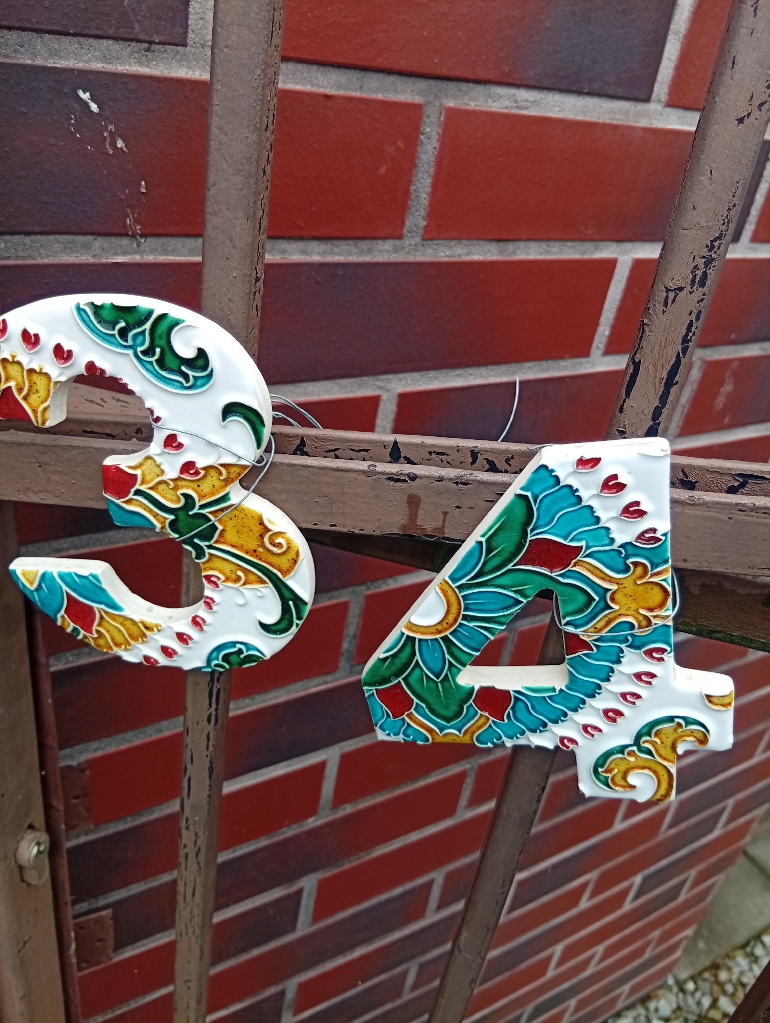 Two decorative house numbers, "3" and "4", are attached to a weathered metal railing. The numbers are white with intricate, colorful designs painted on them, featuring patterns of teal, green, red, and yellow that resemble stylized floral motifs or dragon scales. The "3" is on the left and the "4" is on the right. They appear to be made of ceramic or a similar material. The background is a brick wall with rows of reddish-brown bricks separated by light gray mortar. The metal railing is rusty and has peeling paint, suggesting it is outdoors and has been exposed to the elements. The lighting appears to be natural daylight, casting soft shadows. The image provides a close-up view of the numbers and railing, with the brick wall filling the background. There is no visible text in the image. The overall impression is of a decorative element on an older building's exterior in Wrocław, Poland.