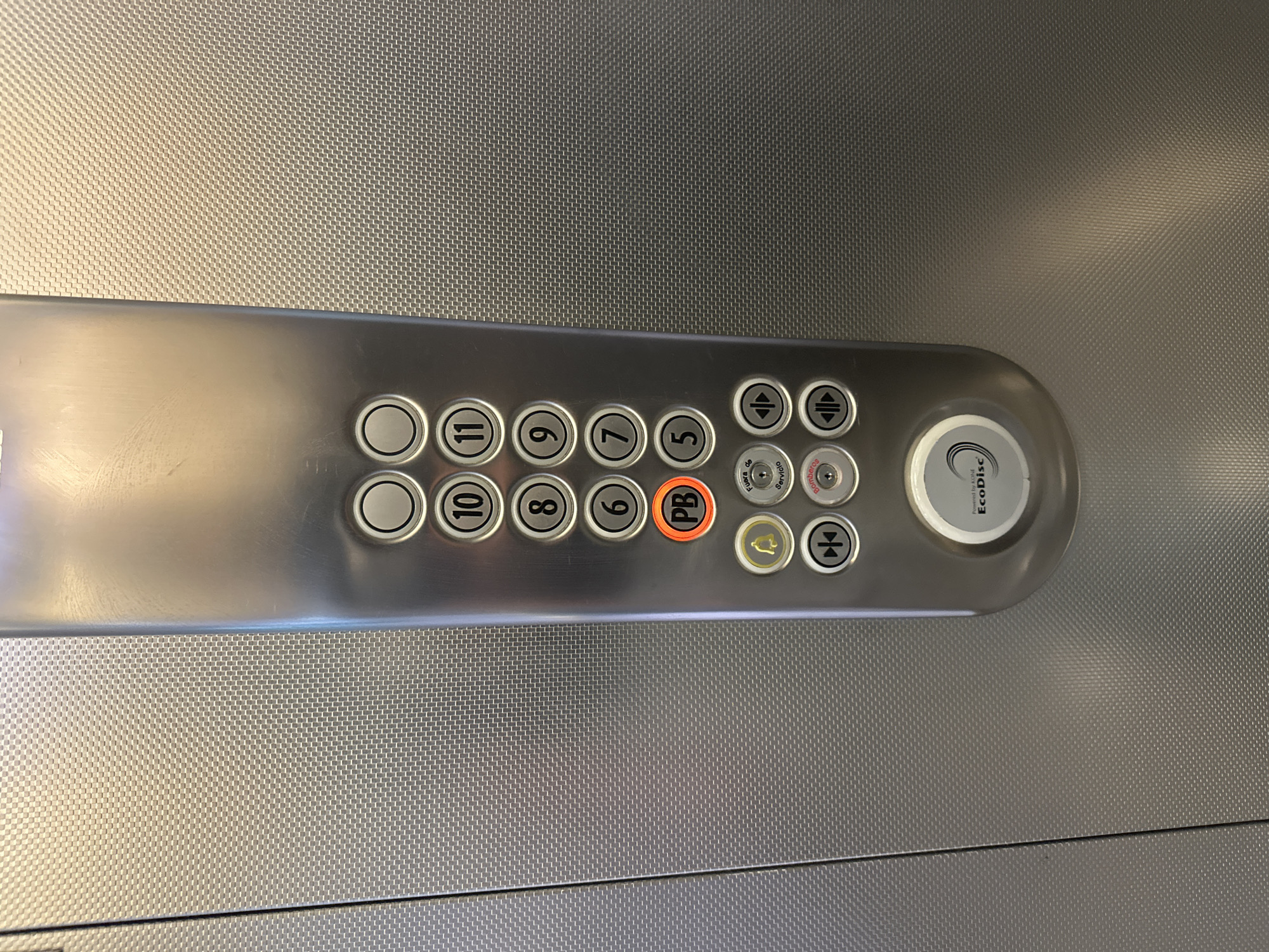 An upward-oriented shot captures an elevator control panel mounted on a textured, metallic wall. The panel is a sleek, brushed metal strip featuring a row of circular buttons for floor selection, ranging from 5 to 11. The numbers visible are 5, 6, 7, 8, 9, 10, and 11, arranged in two rows. A prominent orange-rimmed button labeled "PB" is situated among the floor buttons. To the right of the floor buttons are several smaller, specialized buttons. These include a pair of up and down arrows, a bell icon (likely for an alarm), and horizontal arrows which typically control door opening and closing. Below these, two circular buttons are labeled with text: "Fuera de Servicio" (Out of Service) and "Bombero" (Firefighter), suggesting the elevator is in a Spanish or Portuguese-speaking region. A large, circular badge on the far right of the panel reads "Powered by KONE EcoDisc," identifying the manufacturer and technology of the elevator. The background is a consistent, fine-meshed metallic pattern.