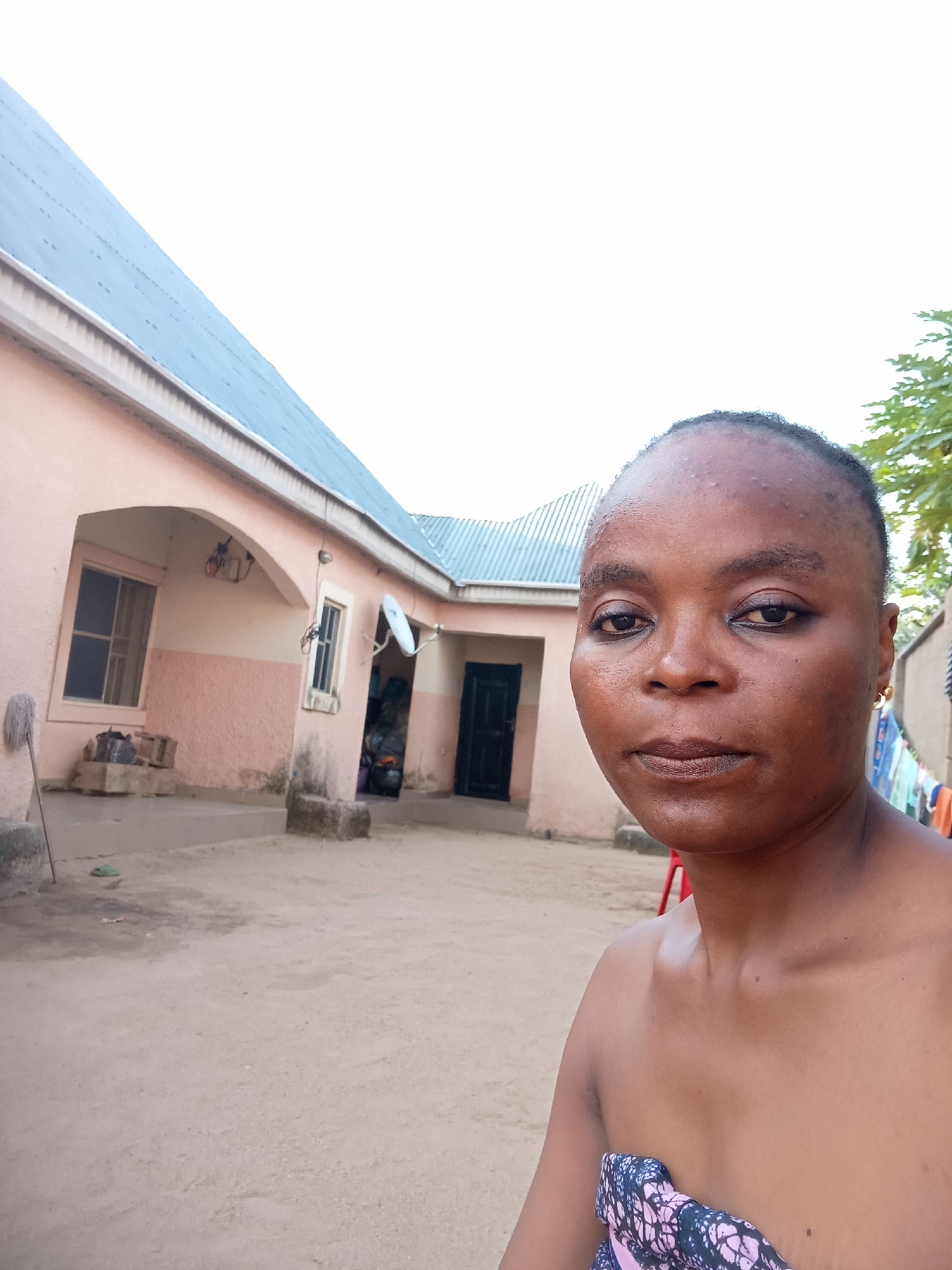 A medium shot selfie is taken from a slightly low angle outdoors in what appears to be a courtyard in Kason, Nigeria. The subject, a woman with dark skin and short, dark hair, is positioned in the right half of the frame, facing the camera with a neutral expression. She is wearing a patterned wrap around her upper chest. Her shoulders and upper chest are visible. Behind her, to the left, is a light pink building with a light blue metal roof. The building has at least two windows, one with a visible grill and another with shutters. An archway with a smaller window is also present. The ground is composed of light brown dirt. To the right of the woman, some laundry is hanging on a line. The sky is overcast and white. The overall scene suggests a domestic setting.