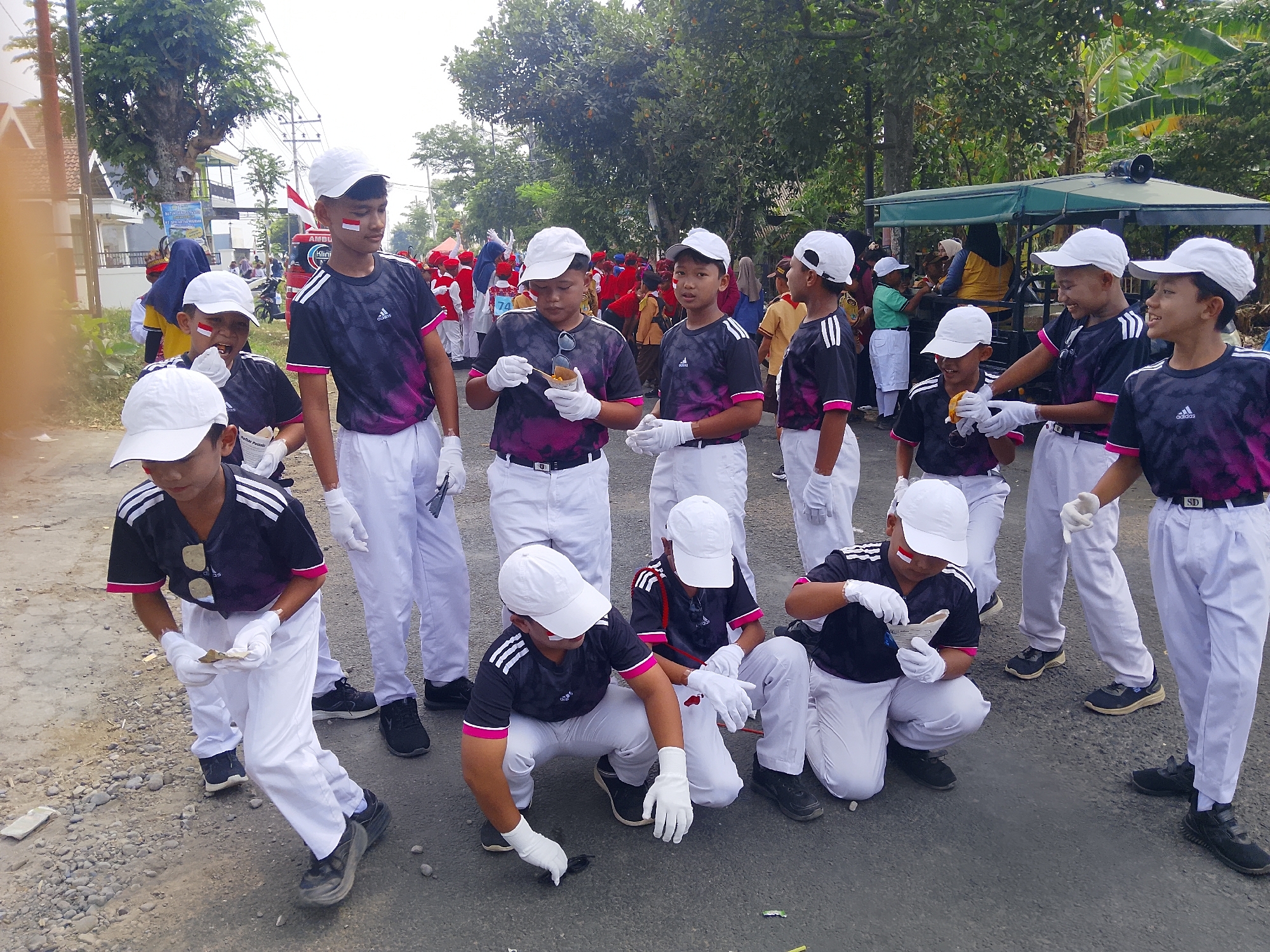 In Bangsri, Indonesia, a group of young boys, likely students, are participating in a parade or event. They are dressed in matching black shirts with pink and white accents, white pants, white caps, and white gloves. Several of the boys have red and white face paint, indicating national pride or participation in a celebratory event. Some boys are holding or interacting with small objects, possibly for a performance or activity. In the background, other participants in red and white attire are visible, along with a green canopy over a cart and trees lining the street. The scene appears to be outdoors on a paved road with some gravel.
