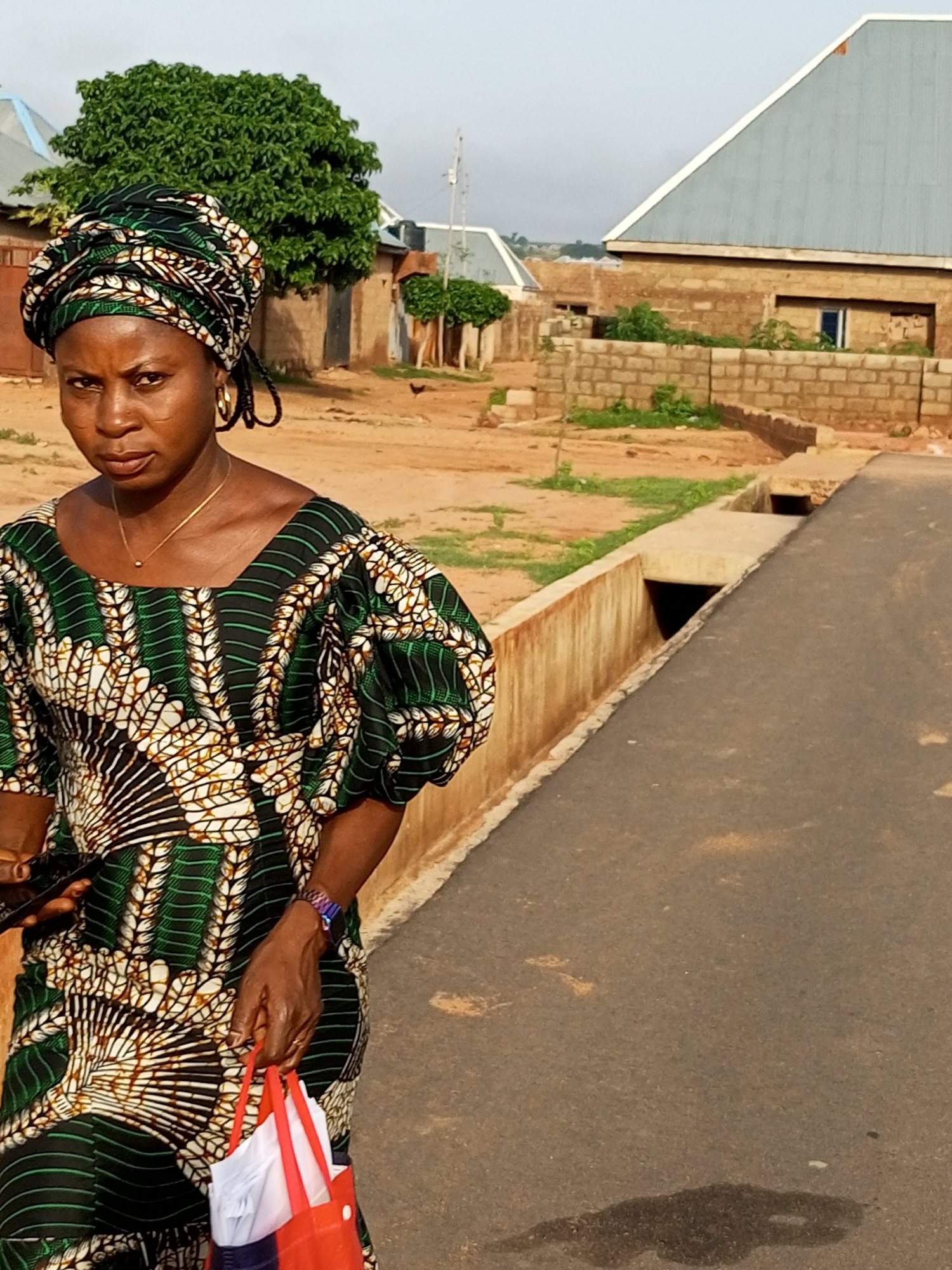 A woman stands on a paved road in what appears to be a developing area of Tempure, Nigeria. She is wearing a colorful, patterned dress and a matching headwrap. A gold chain with a small pendant is visible around her neck, and she has large gold hoop earrings. She is holding a dark-colored mobile phone in her left hand and a red reusable shopping bag in her right. The bag contains white items. A colorful bracelet is on her right wrist. The background shows partially constructed buildings with brick walls and metal roofs, along with some sparse vegetation and a drainage ditch running alongside the road. The ground is a mix of dirt and gravel. The sky is overcast, suggesting it may be early morning or late afternoon.