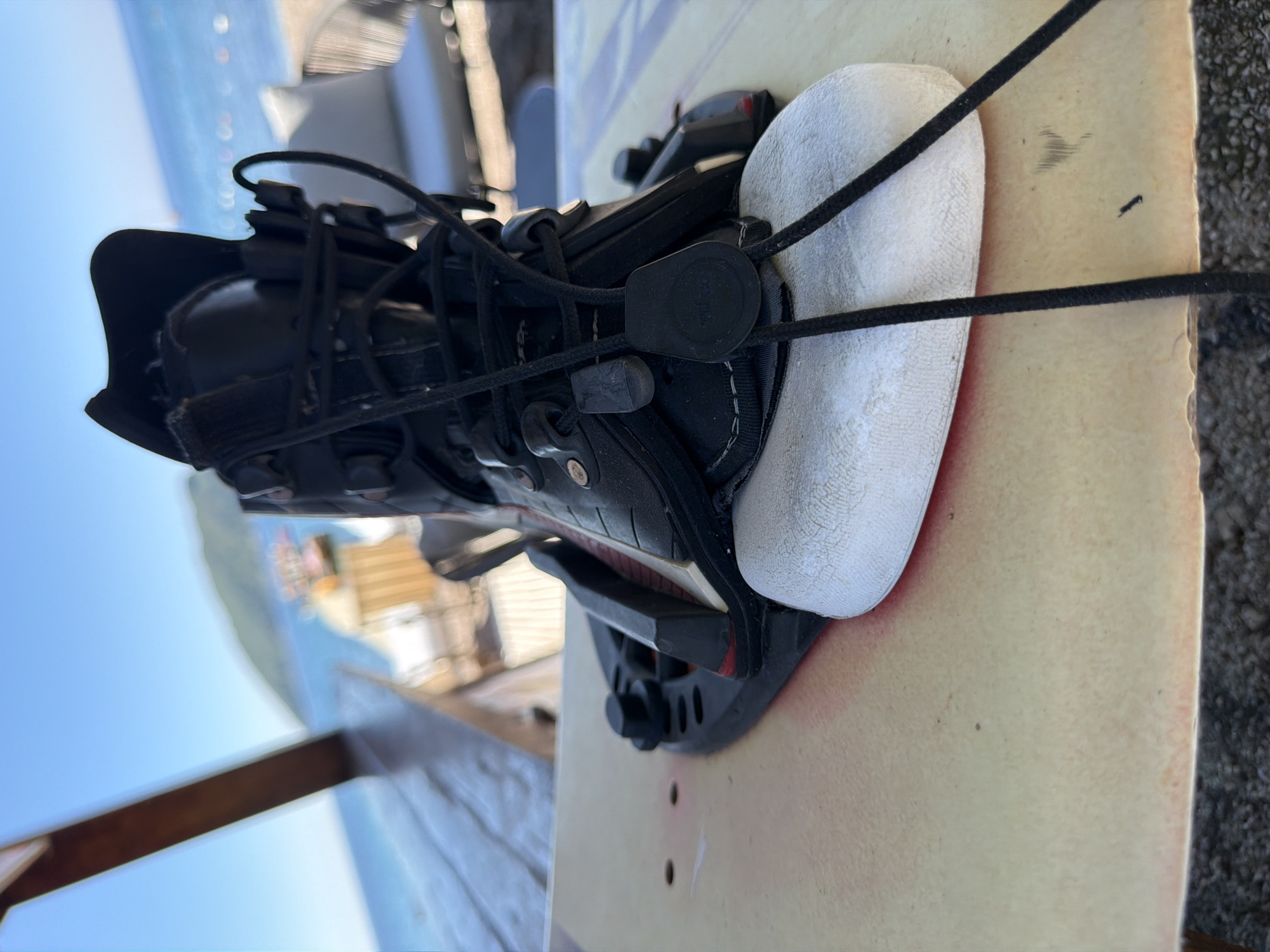 A detailed forensic description of the image follows:
The image shows a close-up of a black wakeboard boot mounted on a light-colored wakeboard. The boot is securely fastened to the board with black bindings. The laces of the boot are black and tightened with a pull-lock mechanism. The interior of the boot features a white padded lining around the ankle area. The wakeboard appears to have a textured surface and is positioned outdoors on a wooden surface.
In the blurred background, there is a body of water visible, possibly the sea. The horizon is partially visible with a light blue sky above. A blurred building is visible in the background as well.
Based on the information available, it is not possible to identify the location as the city of Budva, Montenegro.