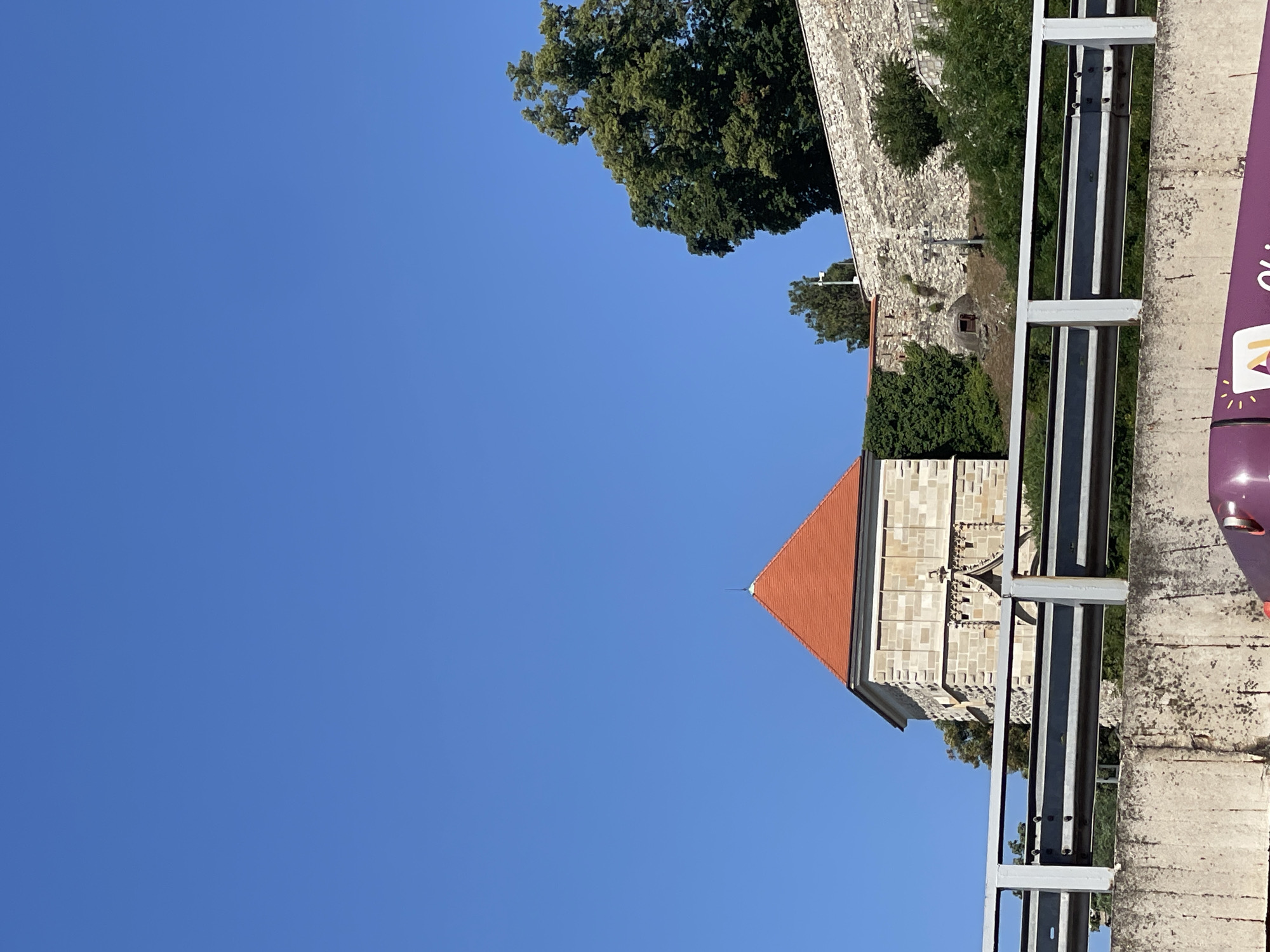 This image shows a portion of Bratislava Castle in Bratislava, Slovakia, viewed from below a concrete overpass with a metal guardrail. In the background, a stone wall of the castle complex runs along a hillside, partially covered by green ivy and foliage. A prominent stone tower with a red-tiled, pyramidal roof is visible on the left side of the castle wall. The sky is clear and blue. The foreground shows the edge of the overpass and a hint of a purple vehicle with a logo.