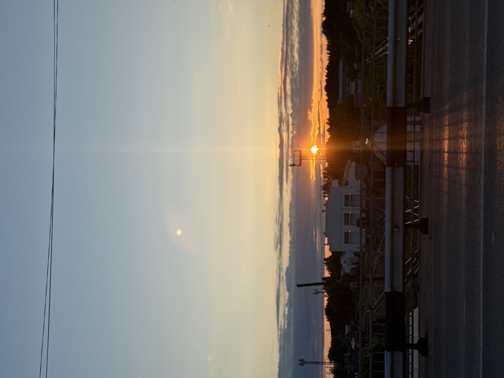 The image captures a vibrant sunset or sunrise over an urban or industrial landscape in Chaikovsky, Russia. The photograph is taken from an elevated position, possibly a bridge or overpass, looking towards the horizon.
The most striking feature is the sun, positioned low on the horizon towards the right side of the frame, emitting a brilliant orange and yellow glow. Strong lens flare emanates from the sun, creating bright streaks of light across the sky. The sky transitions from a light blue at the top, through soft yellows and oranges around the sun, to deeper orange and purplish hues near the horizon, where scattered clouds are visible, illuminated by the sun.
Along the horizon, a dark silhouette of a treeline is visible, interspersed with various buildings. Some structures appear to be large industrial or commercial buildings, with discernible windows and architectural details, silhouetted against the bright sky.
In the foreground on the right, a dark asphalt road with faint lane markings is visible, suggesting a transportation route. A metallic guardrail or barrier runs along the edge of the road, its vertical supports casting long shadows onto the road surface due to the low angle of the sun. On the far left, thin power lines cut across the lighter part of the sky.
The time of day is clearly either dawn or dusk, characterized by the dramatic colors of the sky and the long shadows. No people are visible, and there is no discernible text in the image. The scene conveys a sense of peaceful transition, highlighting the interplay between natural light and human-made environment.