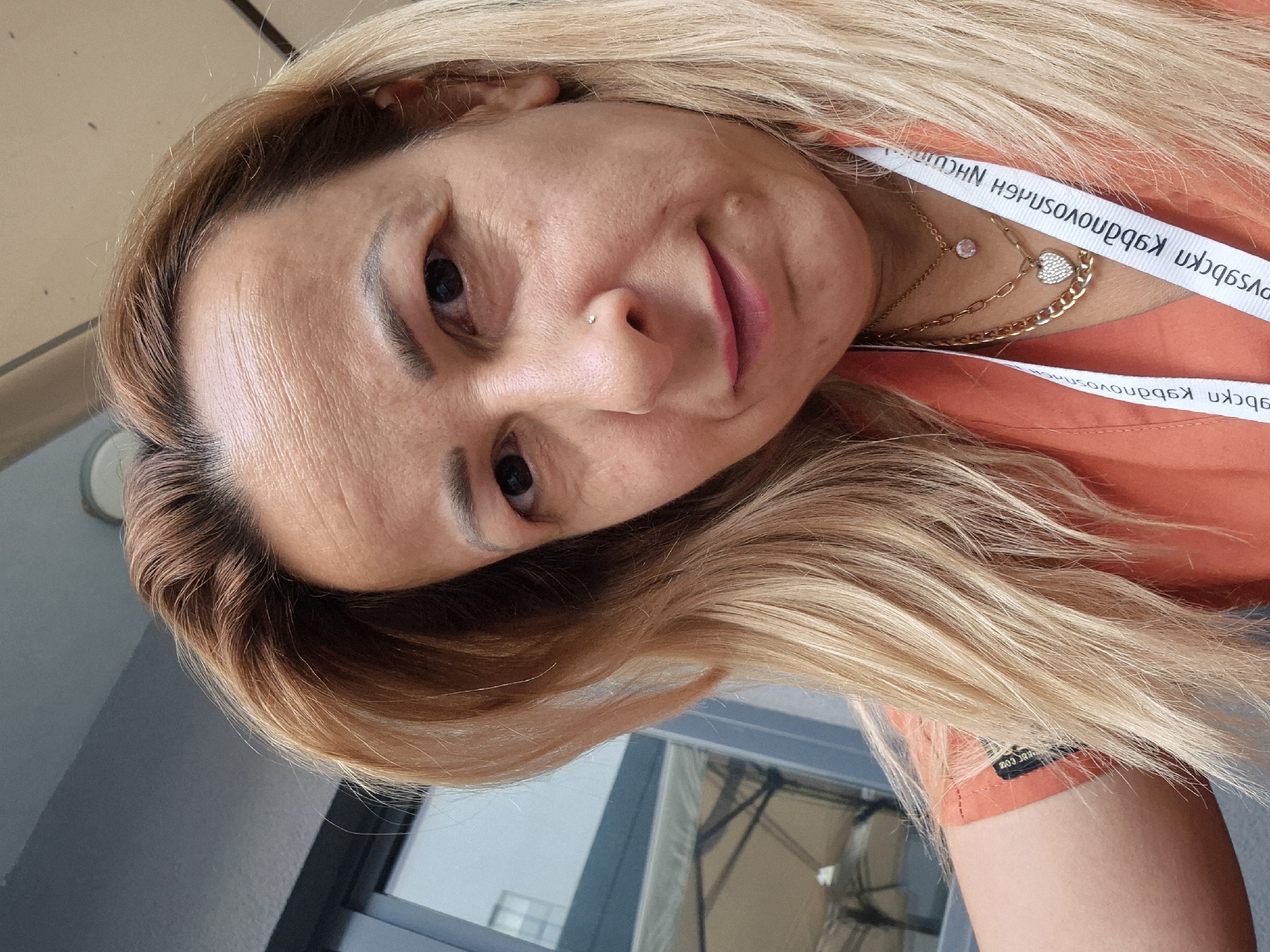 A woman with shoulder-length blonde hair is in an outdoor setting, possibly a cafe or restaurant. She is looking directly at the camera and smiling slightly. The woman is wearing an orange shirt and several necklaces, including one with a heart-shaped pendant. She also has a nose stud. In the background, there are reflections of umbrellas and chairs in a window.