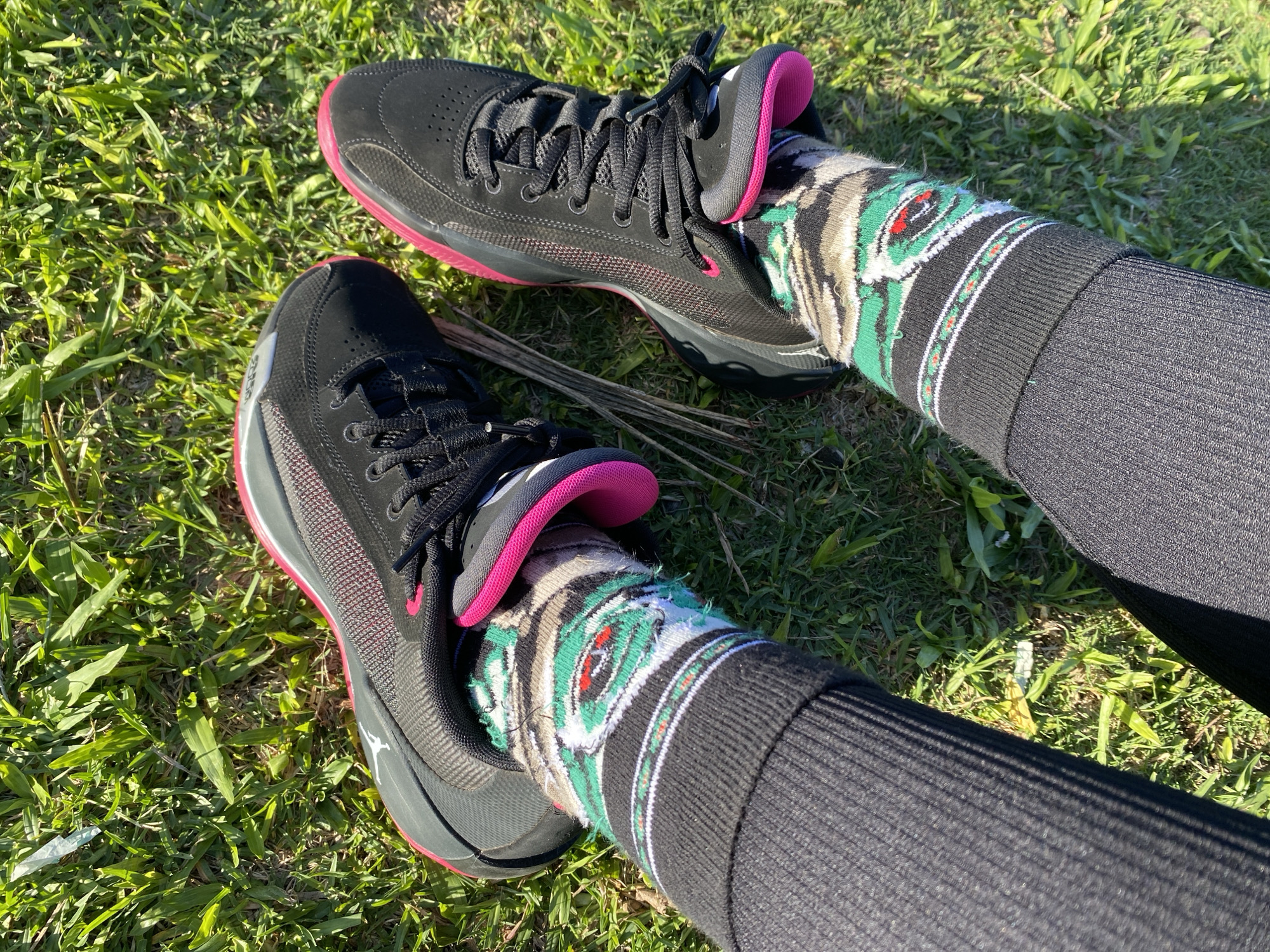 The image shows a person sitting on grass, wearing black leggings, socks with a Southwestern-inspired pattern, and black athletic shoes with pink accents. The location is grass, but the city of Buenos Aires, Argentina, is not discernible. The subject is the person wearing the shoes and socks, and the objects are the leggings, socks, shoes, and the grass. The action is sitting.