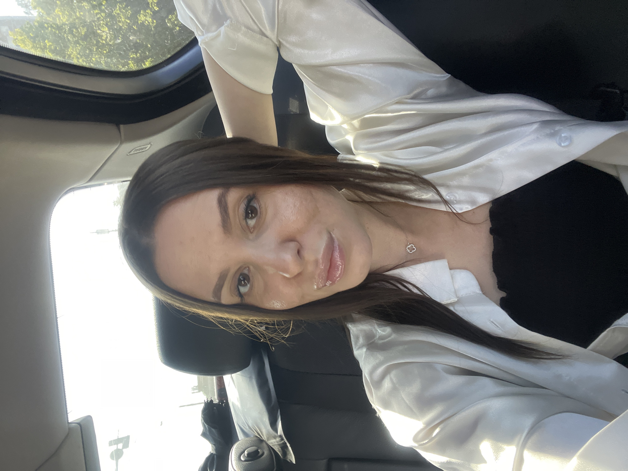 A woman is photographed in the interior of a car. The image appears to be a selfie, with the woman facing the camera. She has long, dark hair and is wearing a white satin shirt over a black top. A silver necklace with a clover-shaped pendant is visible around her neck. The car interior is black, with a view of the outside visible through the window and sunroof. The scene is brightly lit, suggesting daytime. No further information can be determined about a location in Moscow.