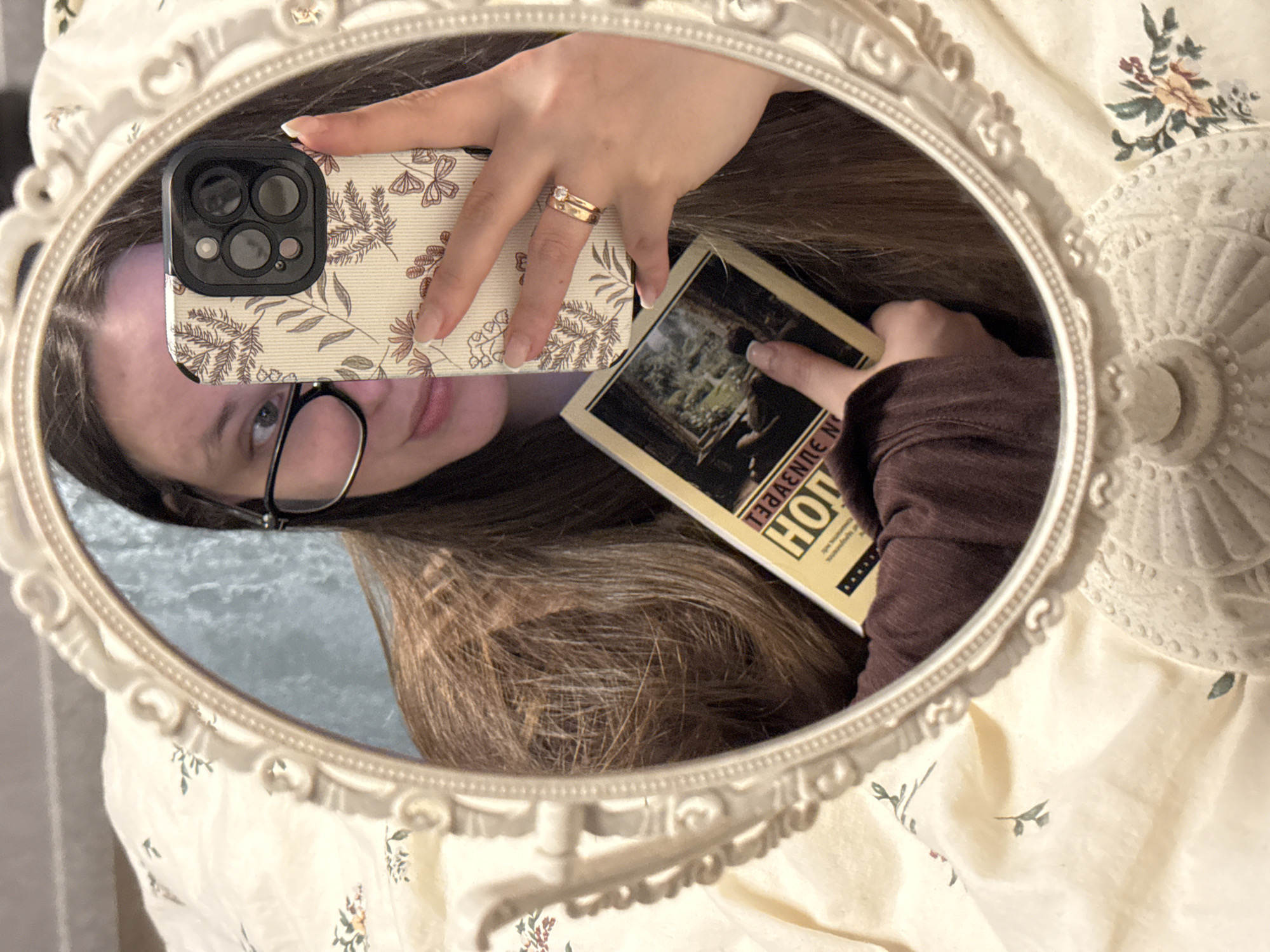 The image depicts a female subject taking a mirror selfie in what appears to be a bedroom in Krasnodar, Russia. The subject, with long brown hair and eyeglasses, is partially visible, holding a smartphone with a floral-patterned case in her right hand (wearing a gold-colored ring with a stone on her ring finger) and a book with a Cyrillic title in her left hand. Her facial expression suggests a slight smile. The smartphone's camera module, featuring multiple lenses, is prominent. The oval-shaped mirror has an ornate, off-white frame with decorative carvings. The bedding surrounding the mirror is light-colored with a scattered floral pattern of small roses and green leaves. A light blue, textured surface is visible in the reflection behind the subject.