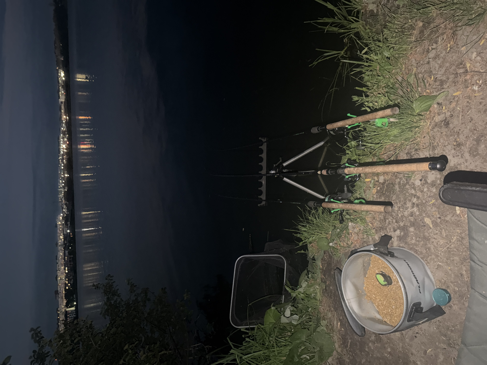 The image shows a nighttime scene at the edge of a body of water, likely a lake or river, with a city skyline illuminated in the distance. In the foreground, a fishing setup is visible. Three fishing rods are propped on a rod pod facing the water. To the left of the rods, a fishing net is placed on the grassy bank. A bucket filled with what appears to be fishing bait is situated in the lower left of the frame, with two green lids nearby. The water is dark and reflects the distant city lights, creating a shimmering effect. The bank is covered in grass and dirt. The overall atmosphere is tranquil and suggests a fishing excursion at dusk or night.