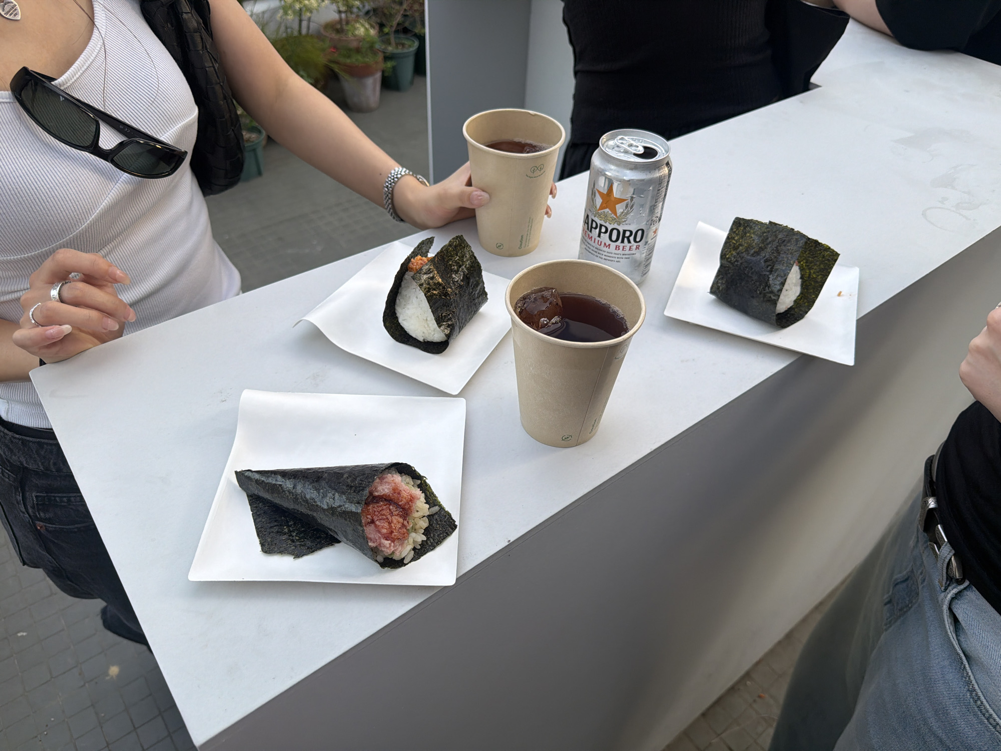 The scene shows two people at a white counter eating and drinking. The individual on the left is wearing a white tank top with sunglasses hanging from the neckline, jeans, and a black shoulder bag. The person on the right is wearing a black top and jeans with a chain belt. On the counter, there are three plates of temaki sushi (seaweed-wrapped rice with fillings), two cups of beverages, and a can of Sapporo beer. The setting is outdoors, with some potted plants visible in the background, suggesting a restaurant or cafe in a city location, potentially in Seoul, South Korea, based on the Sapporo beer which is available in the area, and the type of sushi.