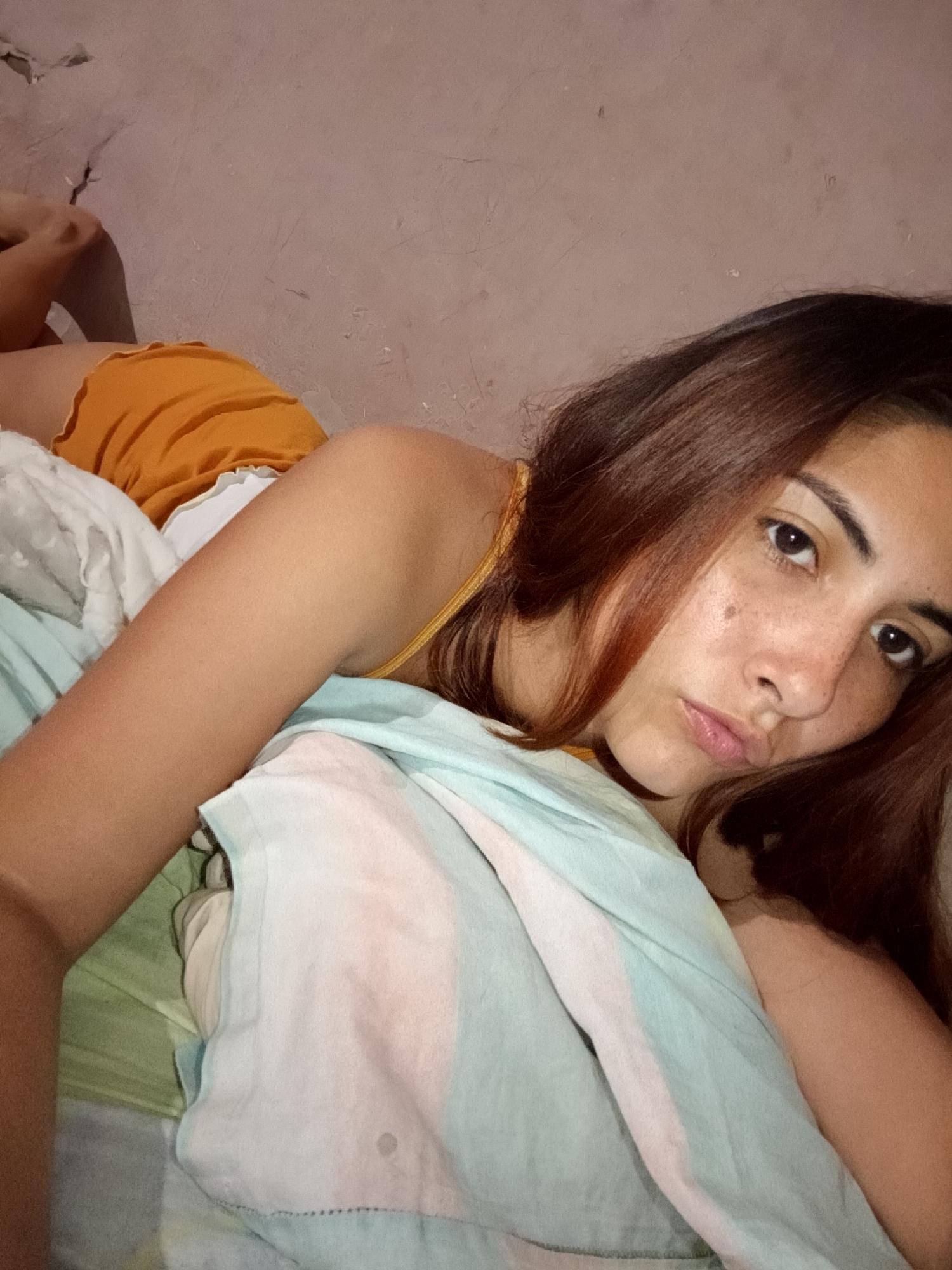 A young woman with brown hair and freckles lies on a bed, propped up on her arm and looking at the camera. She is wearing a yellow top and orange shorts. The background is a bare, textured wall. The bedding includes a white sheet and a light blue and pink striped comforter. The scene appears to be indoors, likely in a bedroom. The lighting is dim, suggesting it may be nighttime or a poorly lit room. The image is a selfie taken from a low angle. The location is specified as Juan Griego, Venezuela.