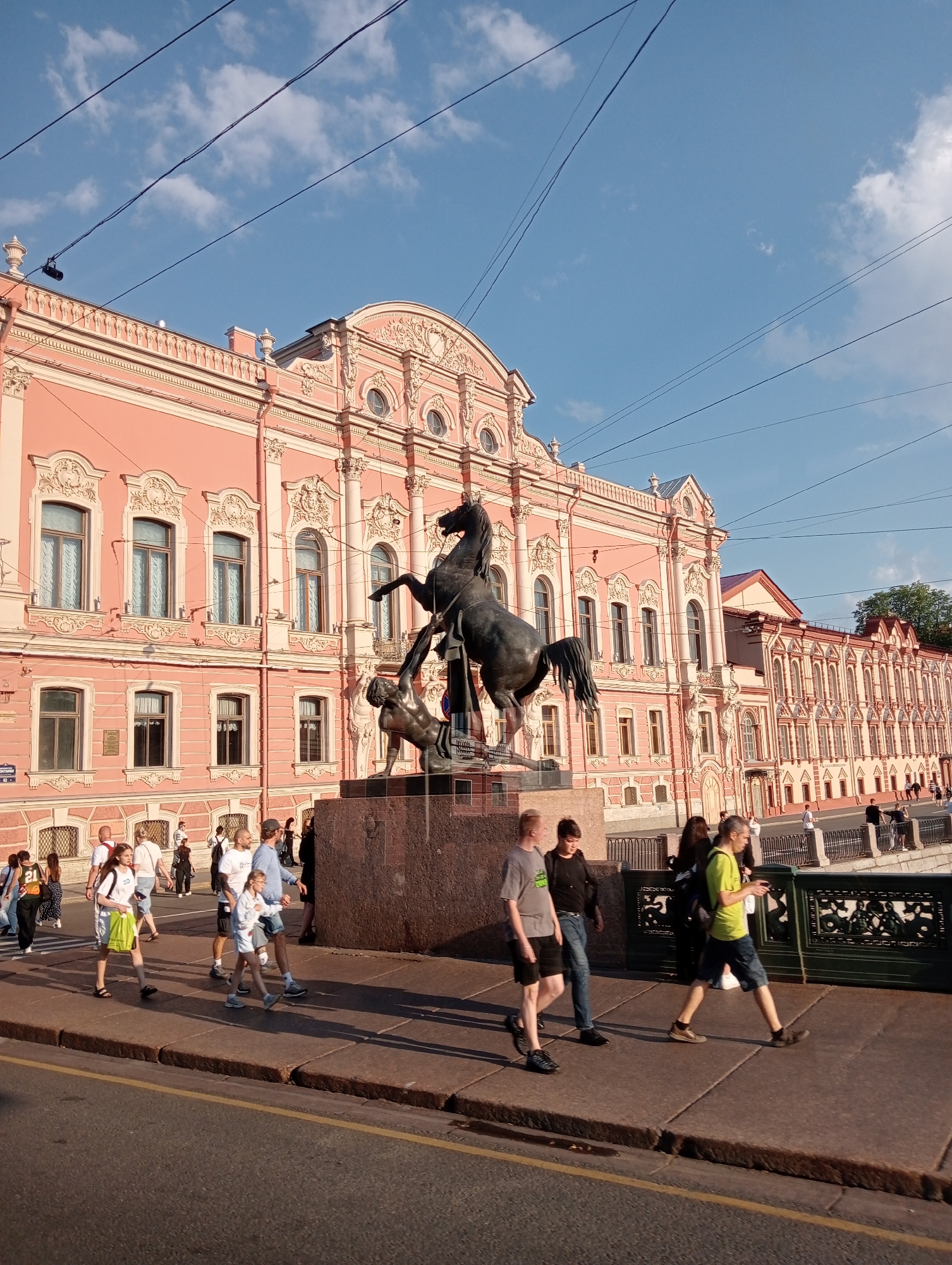 The image depicts a sunny day in Saint Petersburg, Russia, with a clear blue sky dotted with fluffy white clouds. The scene features a grand, pink neoclassical building with ornate white stucco decorations and numerous windows. In the foreground, a bronze equestrian statue stands on a granite pedestal. The statue depicts a horse rearing up on its hind legs, with a figure beneath it. A group of people are walking along a paved walkway in front of the building and the statue. They are casually dressed, suggesting a warm day, and appear to be tourists or locals enjoying an outing. One person is carrying a green bag, and another is holding a phone. The presence of overhead wires suggests an urban environment, possibly with trams or trolleybuses. The architecture of the building and the style of the statue strongly indicate a historical and culturally significant location within Saint Petersburg. The overall atmosphere is peaceful and pleasant, with the sunlight casting shadows on the walkway.