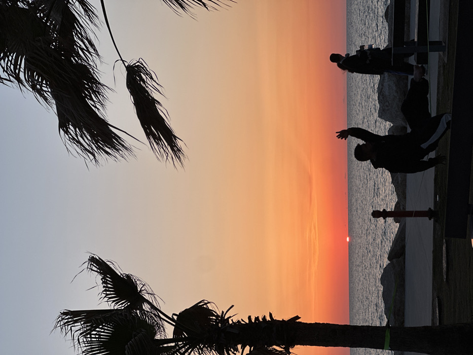 The scene depicts a sunset over the Mediterranean Sea along the Tel Aviv coastline. The sky exhibits a gradient from light blue at the top, transitioning through orange to deep red at the horizon, where the lower portion of the sun is visible as a reddish-orange disc. The sea, appearing dark blue-gray, shows minor surface choppiness.
In the foreground and mid-ground, several elements are silhouetted against the bright sky. A prominent palm tree trunk with dense fronds occupies the far left of the frame; a green band is visible around its lower trunk. Additional palm fronds extend into the upper right corner.
On a concrete promenade running parallel to the sea, a male individual is captured mid-action, leaning significantly backward with his right arm raised and bent, and his legs extended. He appears to be balancing on a low, dark structure, possibly a bench or a slackline anchored to it, which spans the bottom of the frame. He is dressed in dark clothing with a distinct white stripe on his right pant leg. To the right of this individual and further back on the promenade, two additional silhouetted figures are walking, their specific features indistinct. A red, cylindrical bollard with a dark top stands on the promenade to the left of the central balancing figure. A rocky shoreline separates the promenade from the water.