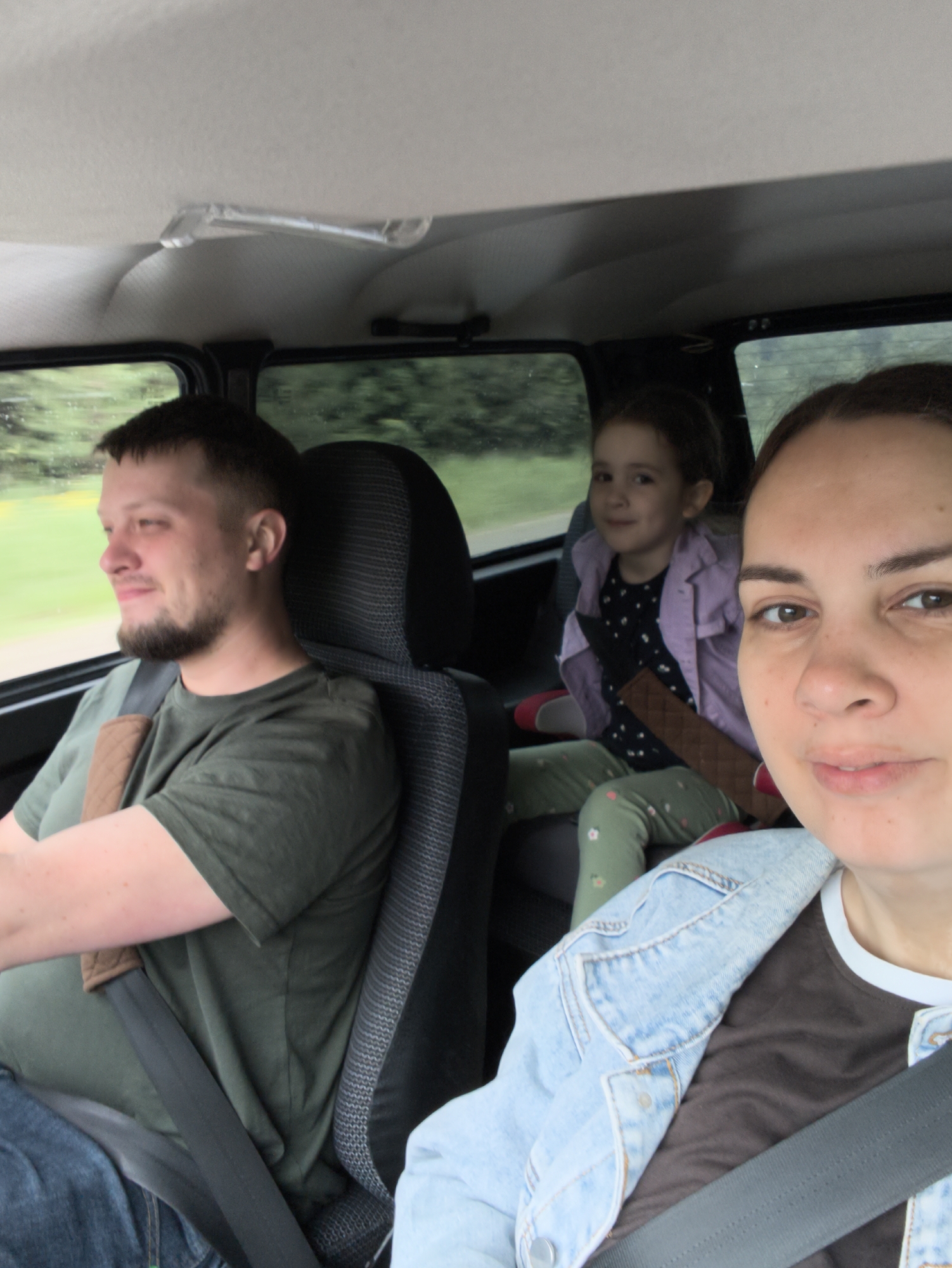 A family of three is captured in a selfie taken inside a car, likely during a daytime journey. The woman taking the photo is in the foreground on the right, partially visible with a neutral expression, wearing a light blue denim jacket over a brown shirt with a white collar. Her seatbelt is visible across her chest.
To her left, a man is driving, seen in profile, smiling softly as he looks ahead or out the window. He has a beard and is wearing a dark green t-shirt and jeans. A brown padded cover is on his seatbelt.
In the backseat, a young girl is visible, positioned slightly behind and to the left of the woman. She is smiling towards the front, seated in a booster seat (parts of red and black are visible). She is dressed in a light purple jacket over a dark top with light patterns, and green pants with small floral patterns. A brown seatbelt pad is also visible across her.
The car's interior appears functional, with a light-colored ceiling and dark fabric seats. Through the car windows, a blurred green landscape of trees and foliage suggests the vehicle is in motion, possibly on a road bordered by nature. The light indicates daytime, and the soft, diffused illumination combined with what looks like moisture on the windows suggests an overcast or slightly rainy day. There is no visible text in the image. The implied setting is a family road trip in Novyi, Russia.