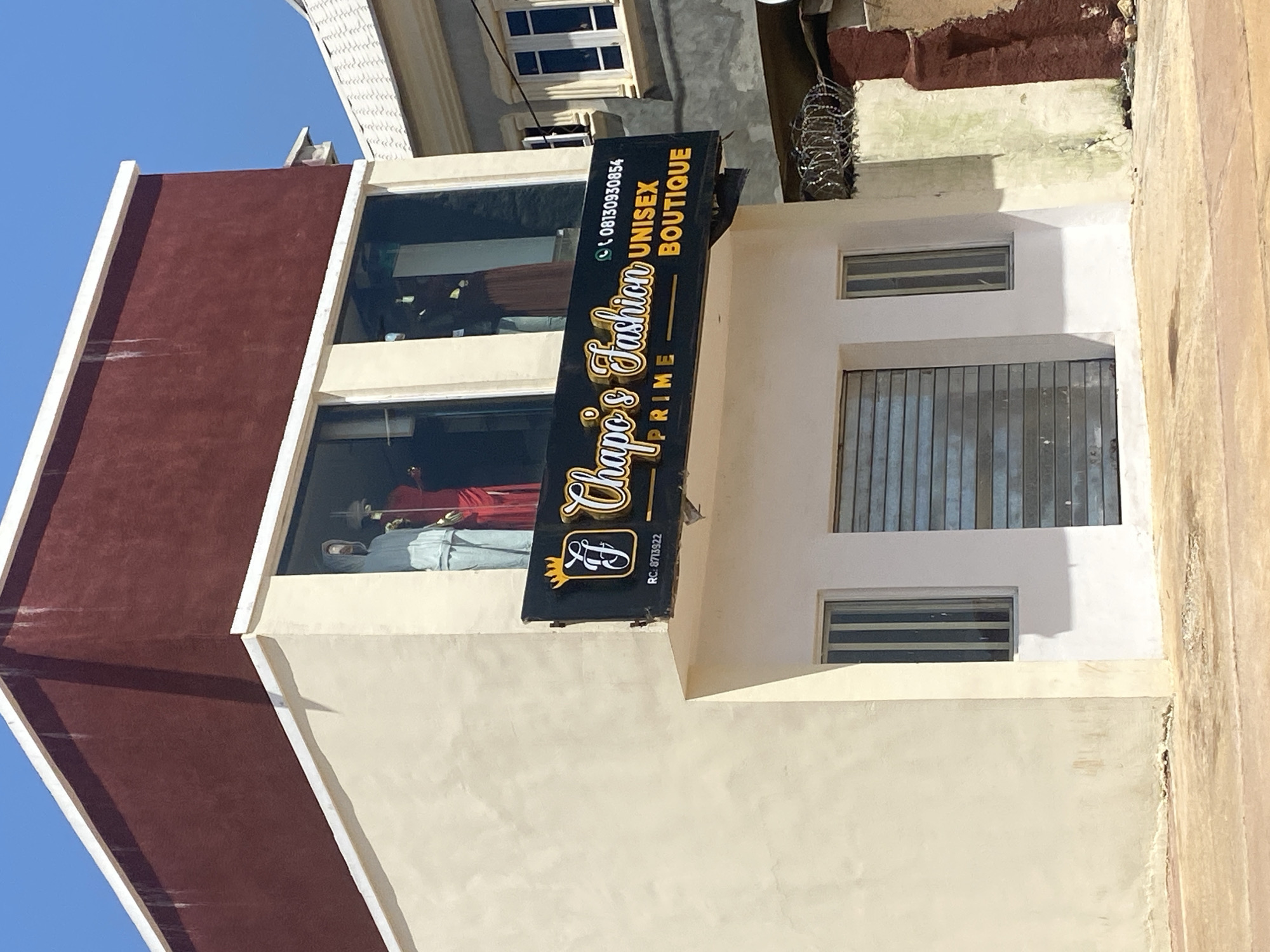 The image shows a two-story building with a maroon upper level and a cream-colored lower level. A sign on the building reads "Chapo's Fashion PRIME UNISEX BOUTIQUE" and includes a phone number. The windows on the upper level display mannequins dressed in clothing. The ground level has a metal shutter and two vertical windows. The building appears to be located in a city. The sky is clear and blue.