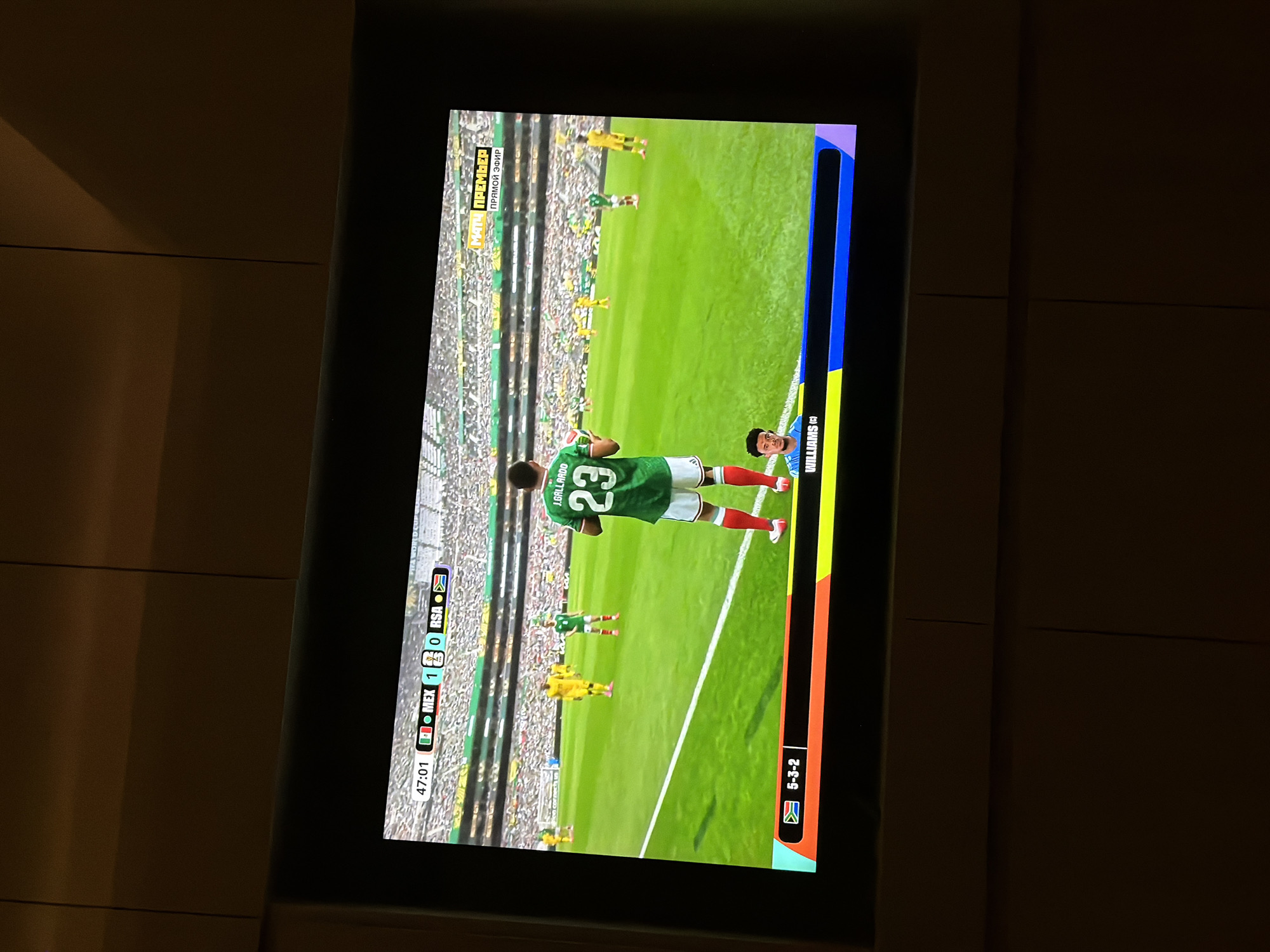 The image shows a television screen displaying a soccer match. The score is Mexico 1, South Africa 0, with the game in the 47th minute. A player in a green jersey with the number 23 is visible on the field. Below the game, a graphic displays the formation "5-3-2" for South Africa. The background shows a stadium filled with spectators. There is a Russian sports channel logo in the top right corner. The scene is taking place in an indoor setting, indicated by the ambient lighting and the nature of the television display.