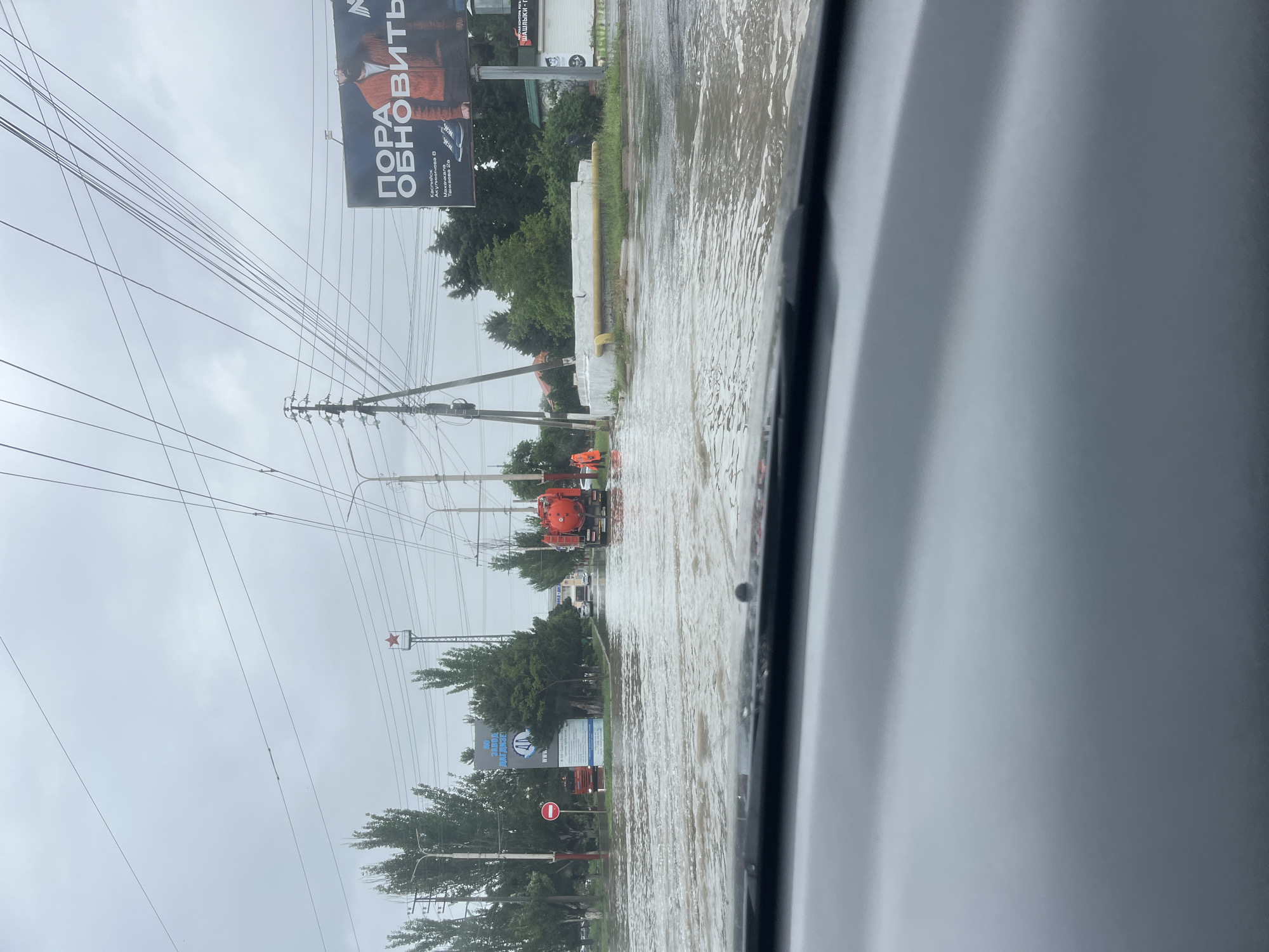 The image shows a flooded street in Kaspiysk, Russia. Water covers the roadway, extending from the sidewalk to an unseen point in the distance. Two orange and red maintenance vehicles are visible in the middle of the flood, appearing to be working on the issue. Overhead power lines stretch across the sky, supported by utility poles. A billboard advertising clothing can be seen on the right. Trees and other infrastructure line the sides of the street. The photograph was taken from inside a car.